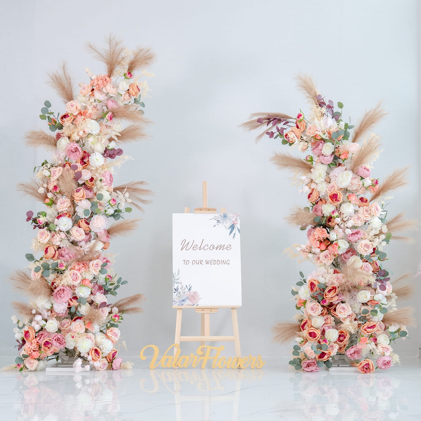 This backdrop features two floral arrangements on either side of a welcome sign that reads "Welcome to our wedding." The flowers include roses in various shades of pink and white, along with eucalyptus leaves and pampas grass. 

The color palette encompasses soft pinks, whites, and hints of green, with the neutral tones of the pampas grass adding a warm contrast. The overall style is romantic and elegant, perfect for a wedding setting, creating a welcoming and dreamy atmosphere. 