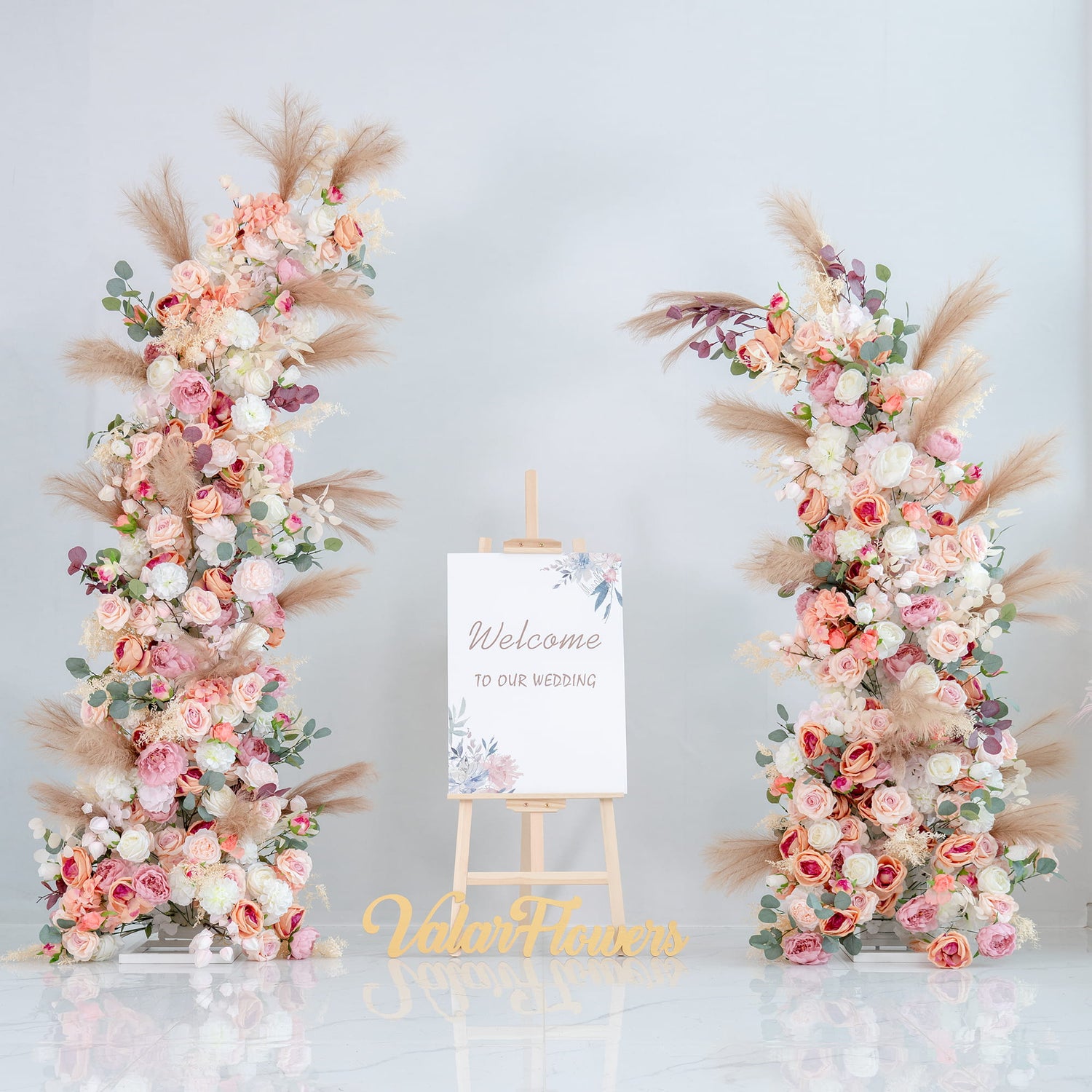 This backdrop features two floral arrangements on either side of a welcome sign that reads &quot;Welcome to our wedding.&quot; The flowers include roses in various shades of pink and white, along with eucalyptus leaves and pampas grass. 

The color palette encompasses soft pinks, whites, and hints of green, with the neutral tones of the pampas grass adding a warm contrast. The overall style is romantic and elegant, perfect for a wedding setting, creating a welcoming and dreamy atmosphere. 