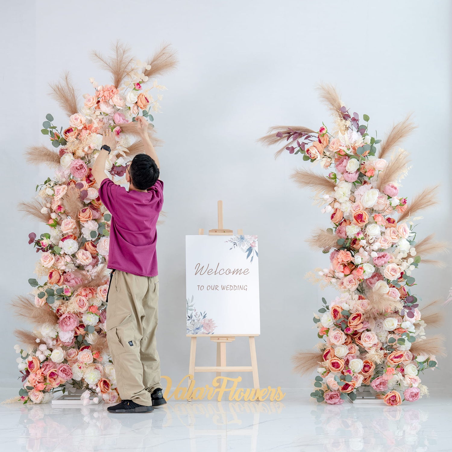 This backdrop features two tall, lush floral arrangements on either side of a wooden easel holding a “Welcome to our wedding” sign. The flowers include roses in various pastel shades, along with peonies, eucalyptus, and pampas grass. The overall style is romantic and elegant, perfect for a wedding setting. 