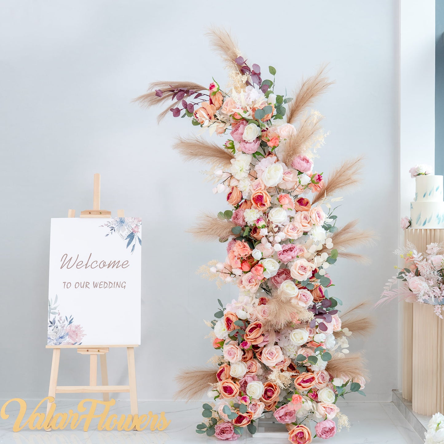 This backdrop features a tall, lush floral arrangement next to an easel displaying a “Welcome to our wedding” sign. The flowers include roses in various shades of pink, white, and peach, along with peonies, eucalyptus leaves, and pampas grass.

The color palette consists of soft pastels like pink, peach, and white, complemented by green foliage and neutral - toned pampas grass. The style is romantic and elegant, perfect for a wedding setting, creating a warm and inviting atmosphere. 