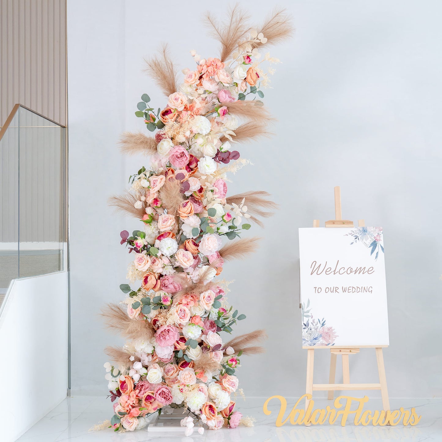 This backdrop features a tall, lush floral arrangement beside a welcome sign that reads "Welcome to our wedding." The flowers include roses in shades of pink, white, and peach, along with eucalyptus leaves and pampas grass. 

The color palette is soft and romantic, with pastel pinks, whites, and hints of green, complemented by the neutral tones of the pampas grass. The overall style is elegant and inviting, perfect for a wedding, creating a warm and dreamy atmosphere. 