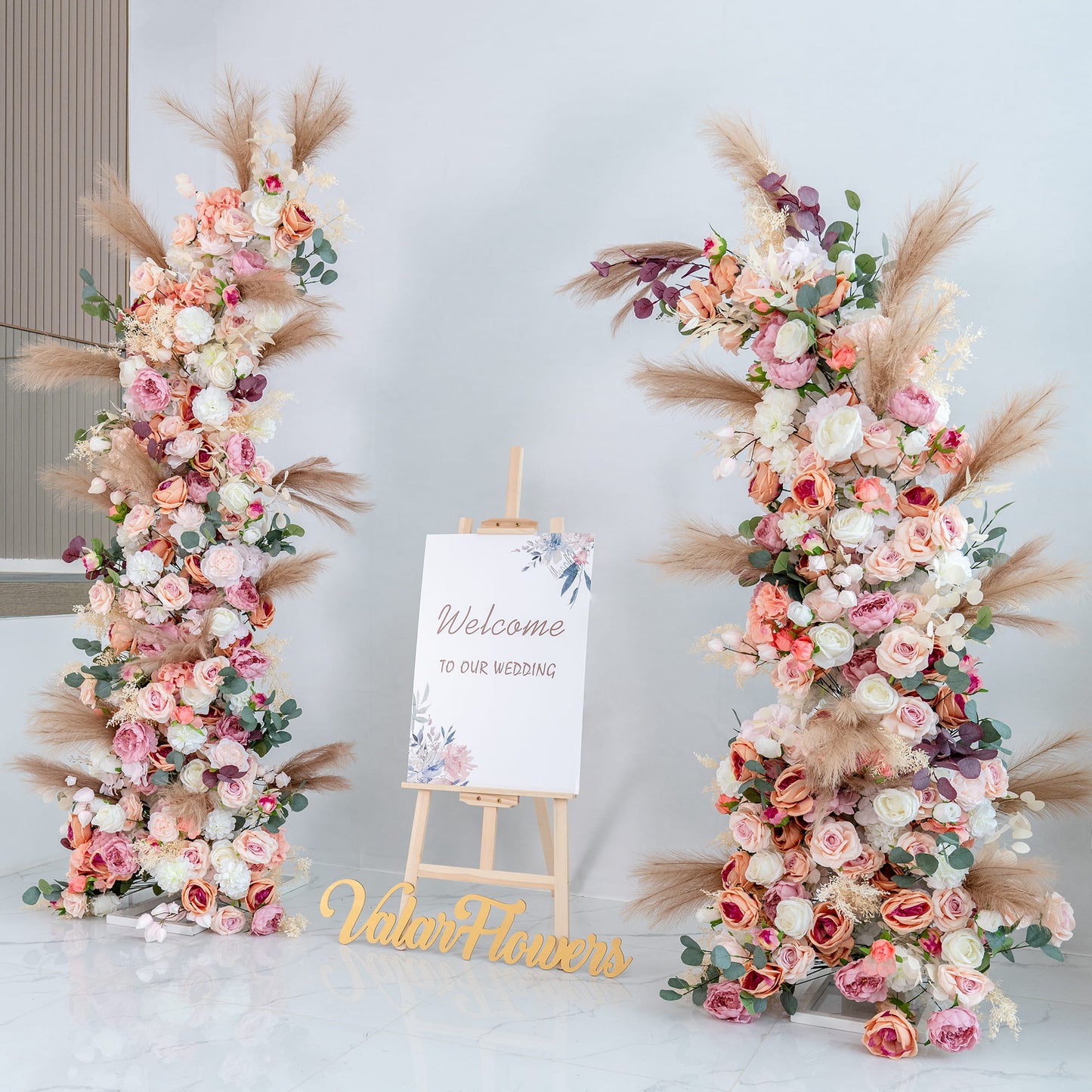 This backdrop features two lush floral arrangements flanking a sign that reads "Welcome to our wedding." Flowers include roses in shades of pink, peach, and white, along with peonies, eucalyptus, and pampas grass. 

The color palette is soft and pastel - toned, with pinks, peaches, whites, and touches of green and beige. The style is romantic and elegant, perfect for a wedding, creating a warm and inviting atmosphere. 