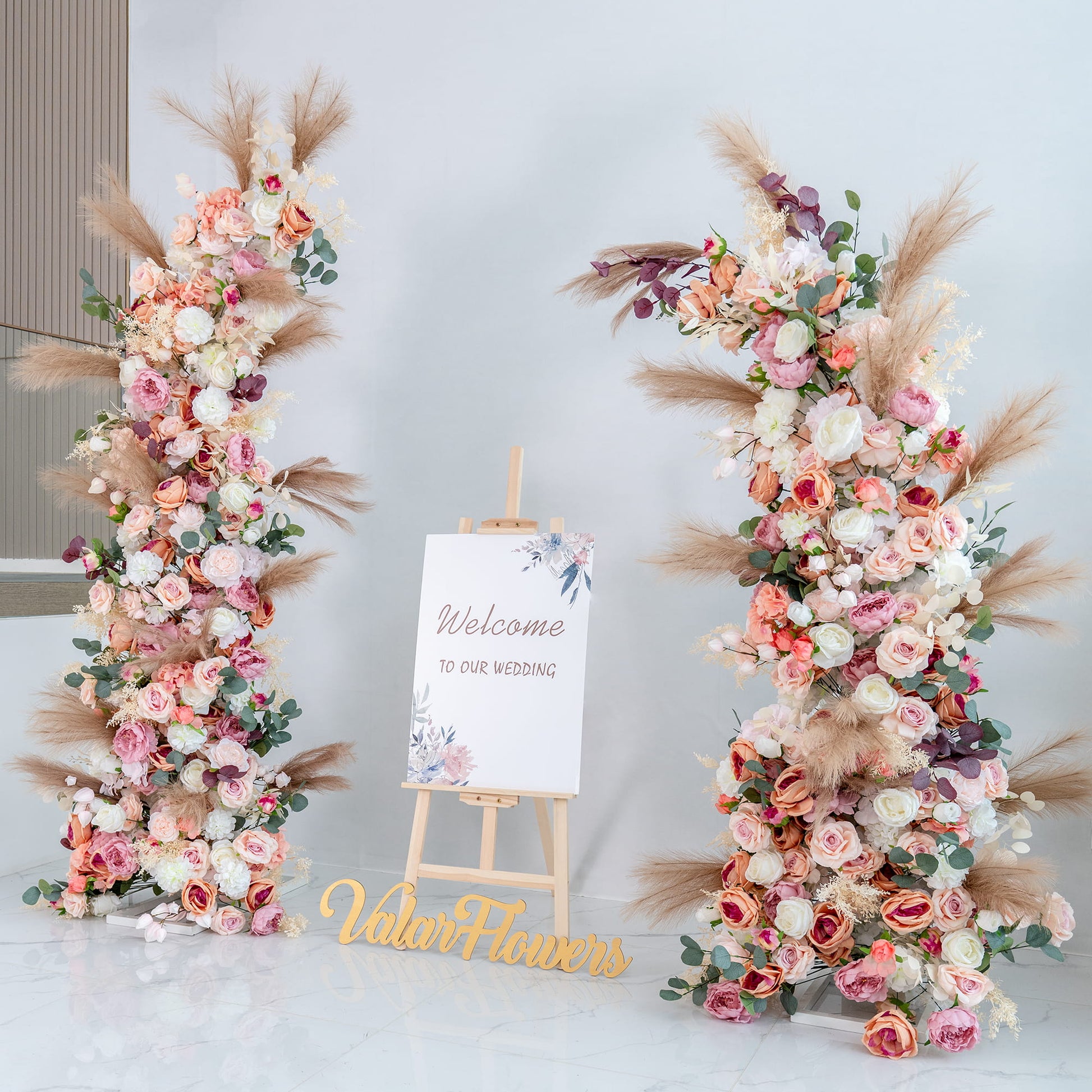 This backdrop features two lush floral arrangements flanking a sign that reads "Welcome to our wedding." Flowers include roses in shades of pink, peach, and white, along with peonies, eucalyptus, and pampas grass. 

The color palette is soft and pastel - toned, with pinks, peaches, whites, and touches of green and beige. The style is romantic and elegant, perfect for a wedding, creating a warm and inviting atmosphere. 