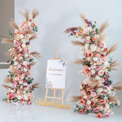 This backdrop features two lush floral arrangements flanking a sign that reads &quot;Welcome to our wedding.&quot; Flowers include roses in shades of pink, peach, and white, along with peonies, eucalyptus, and pampas grass. 

The color palette is soft and pastel - toned, with pinks, peaches, whites, and touches of green and beige. The style is romantic and elegant, perfect for a wedding, creating a warm and inviting atmosphere. 