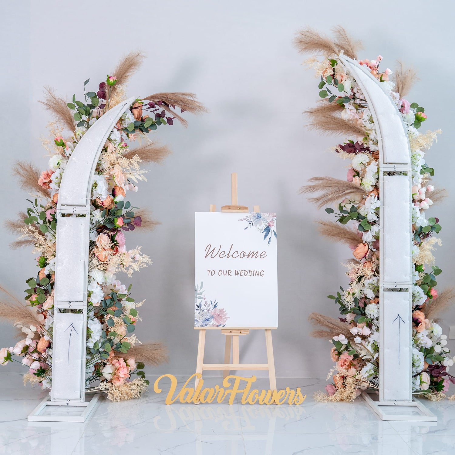 This wedding - themed backdrop features two white - arched floral arrangements adorned with an array of artificial flowers in soft pastel hues, including pinks, peaches, and whites. Pampas grass and greenery add texture. A wooden easel displays a “Welcome to our wedding” sign, creating a warm and inviting atmosphere. 