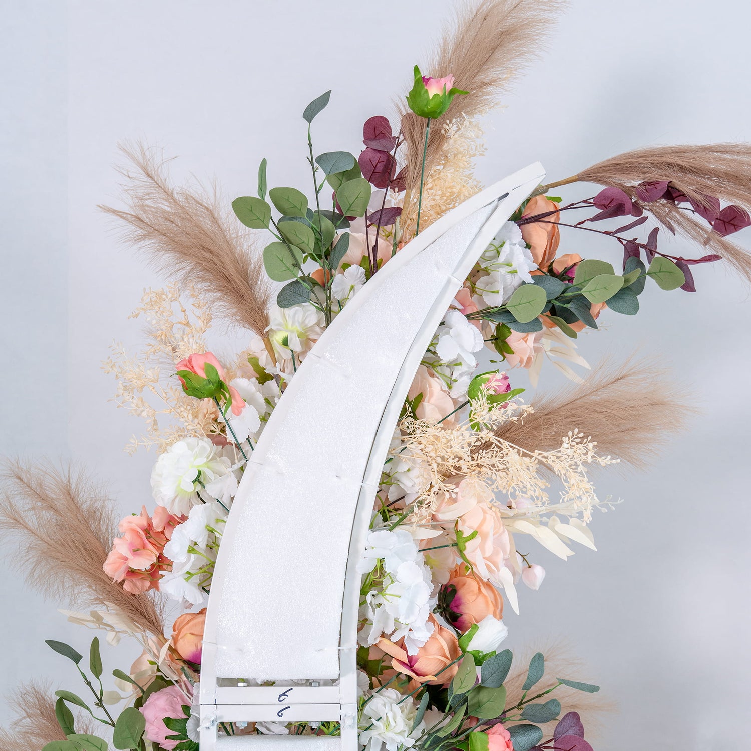 This backdrop features a white, curved structure adorned with an abundance of artificial flowers in soft pastel colors, including pink, peach, and white roses and peonies. Interspersed with green eucalyptus leaves and beige pampas - like feathers, it creates a romantic and elegant atmosphere. 