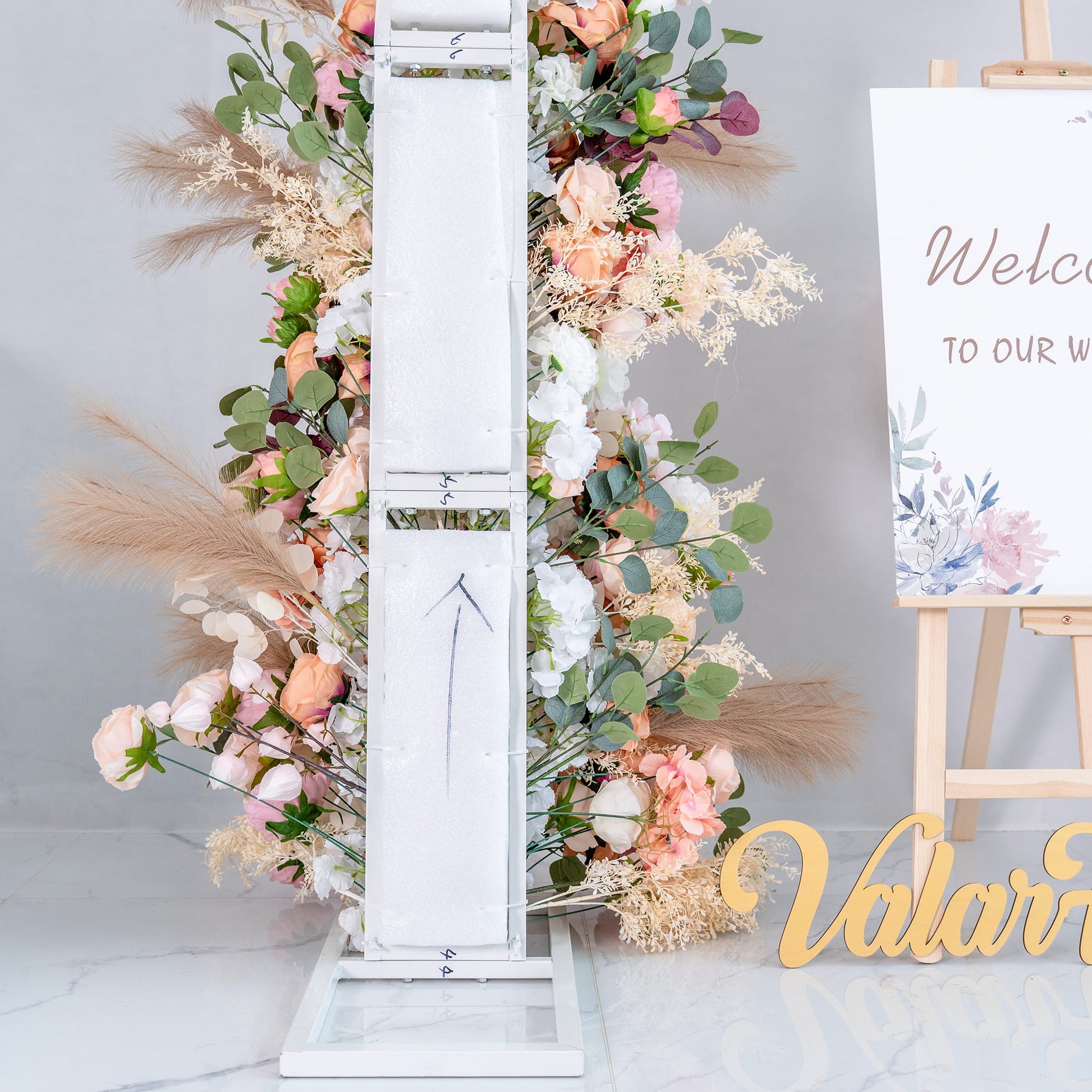 This backdrop features a white structural element adorned with a lush arrangement of artificial flowers. There are roses in various pastel shades, along with peonies and greenery like eucalyptus. Pampas - like feathers add texture. It's part of a wedding setup, with a “Welcome to our wedding” sign nearby. 
