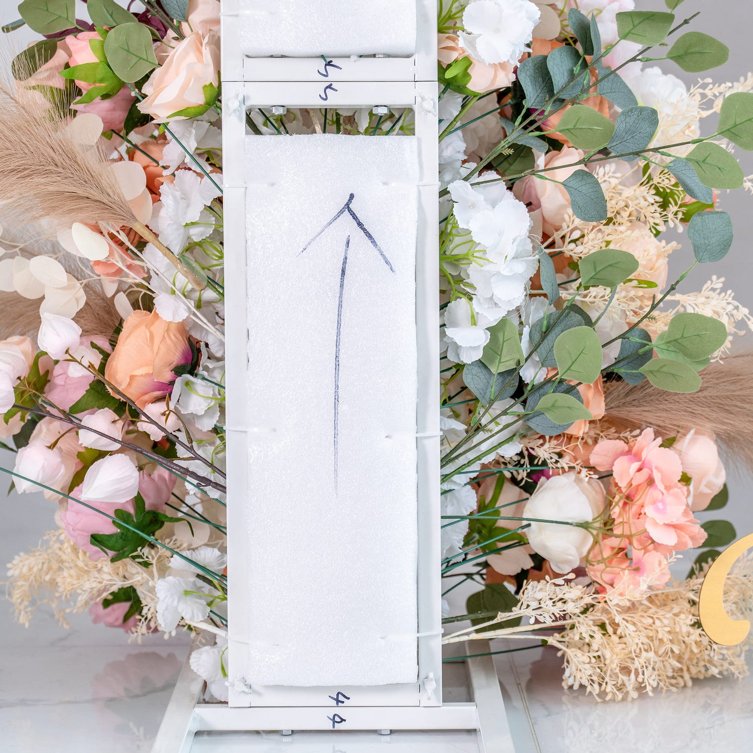 This image shows a white, modular structure with arrows marked on it, surrounded by a vibrant floral arrangement. The flowers include roses and other blooms in shades of pink, peach, and white, along with green eucalyptus leaves and beige pampas - like feathers. The overall scene appears to be part of a wedding or event setup, with a “Welcome to our wedding” sign partially visible in the background. 