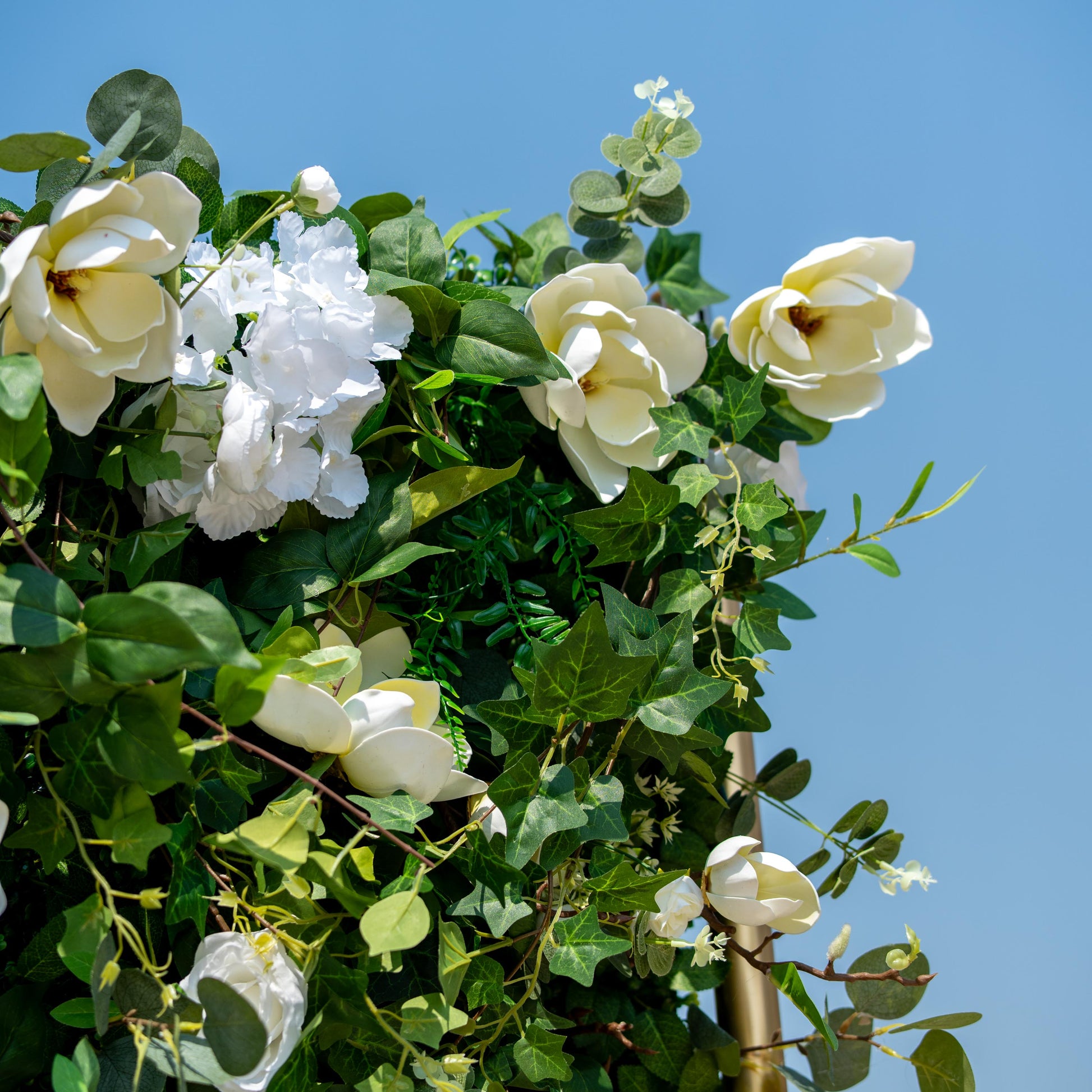 This backdrop showcases a lush arrangement of artificial greenery interspersed with white flowers. Prominent flowers include large, magnolia - like blooms, delicate white roses, and clusters of white hydrangeas. 

The greenery features a mix of eucalyptus - style leaves and various types of ivy - like foliage. The color palette of green and white creates a fresh, natural, and elegant style, ideal for events seeking a serene and sophisticated ambiance. 