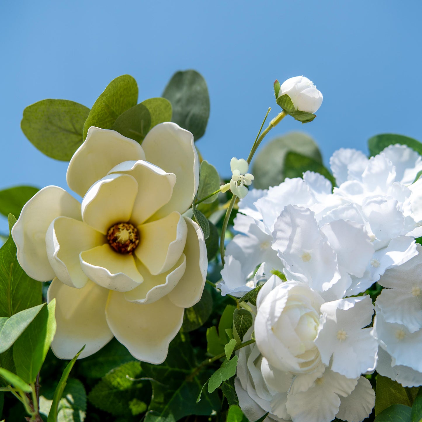 This backdrop features a charming arrangement of artificial flowers and foliage. The flowers include large, creamy - yellow magnolia - like blooms, small white roses, and clusters of white hydrangea - like flowers. The greenery consists of various leaves, such as round eucalyptus - like leaves and pointed ivy - like leaves.

The color scheme is a fresh combination of vibrant green foliage and pure white and creamy - yellow flowers. The overall style is natural and elegant, making it perfect for events like 