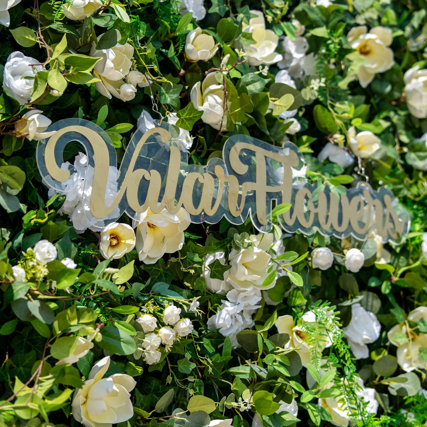 This backdrop showcases a lush floral wall with artificial greenery and white flowers. It features magnolia - like blooms, white roses, and hydrangea - like clusters. The colors are predominantly green and white. 

A sign with “Valar Flowers” adds an elegant touch. The style is natural and sophisticated, ideal for events seeking a fresh, garden - inspired aesthetic. 