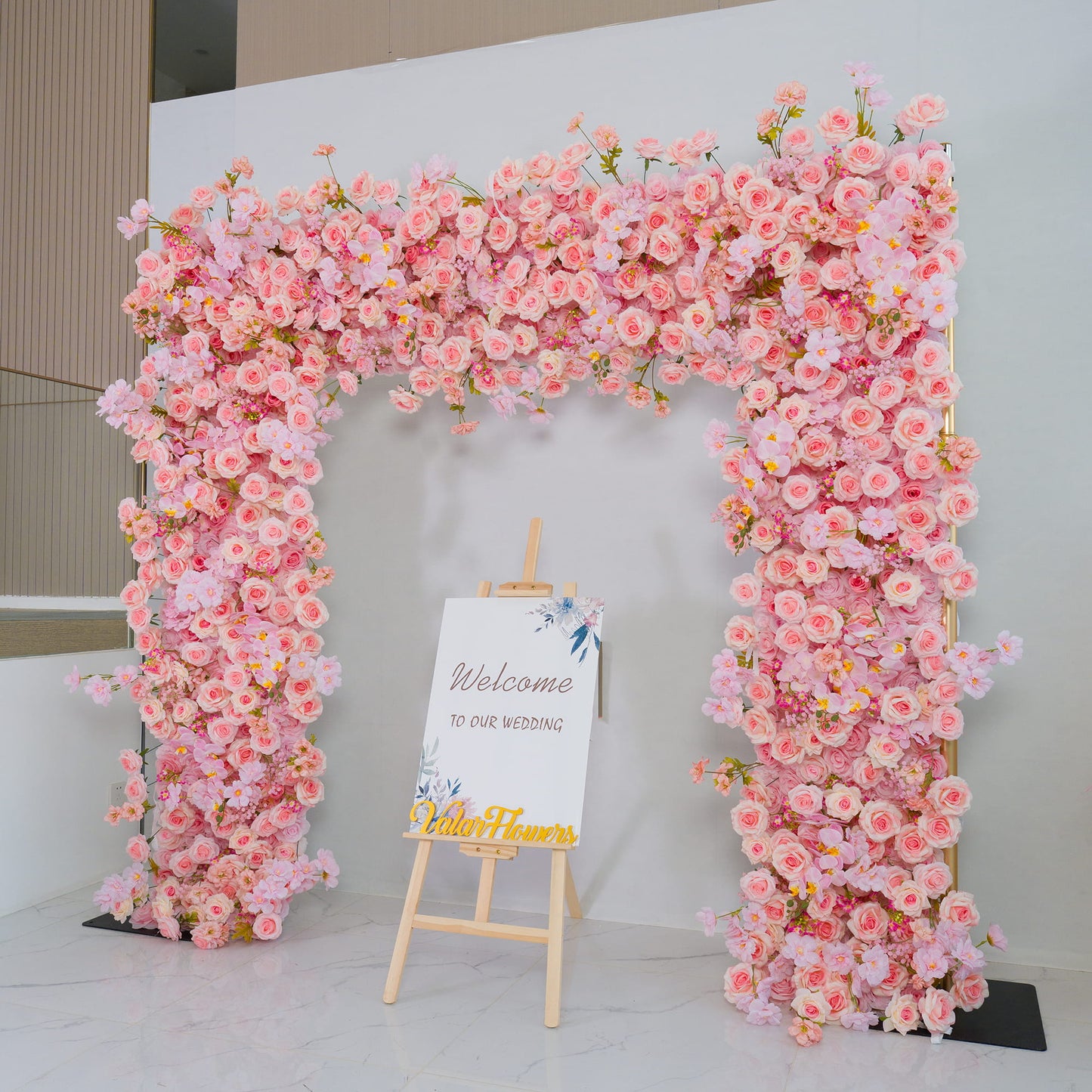 This backdrop is a magnificent floral archway densely packed with artificial pink roses and other blossoms in soft hues. The flowers create a romantic and inviting atmosphere. Below the arch, an easel holds a sign that reads "Welcome to our wedding," enhancing the celebratory mood. Golden - colored text "Valar Flowers" adds a touch of elegance. It is perfect for wedding entrances, creating a charming and memorable first impression. 