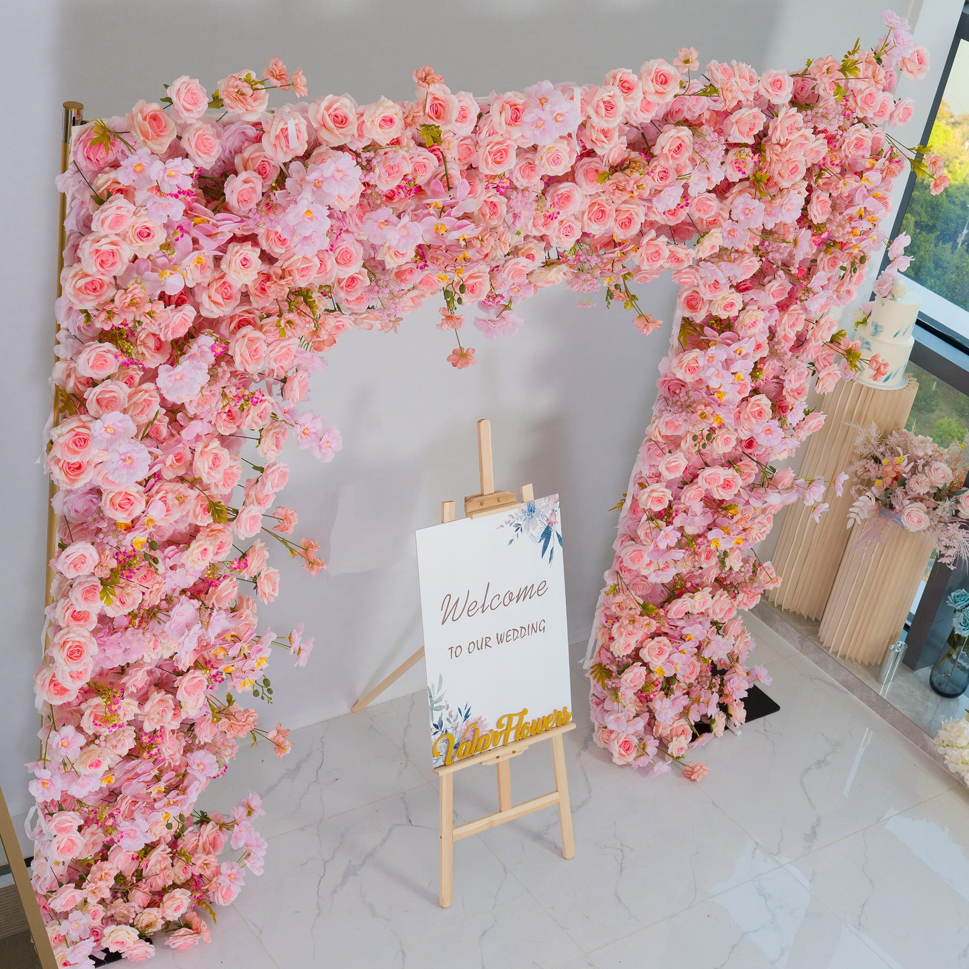 This backdrop is a floral archway adorned with a profusion of artificial flowers. Predominantly in various shades of pink, it features delicate pink roses, large - petaled orchid - like blooms, and small clusters of vibrant pink and pastel - pink filler flowers.

The golden - hued metal poles add an elegant touch. The overall style is romantic and celebratory, perfect for weddings, as indicated by the "Welcome to our wedding" sign. It creates a warm and inviting atmosphere. 