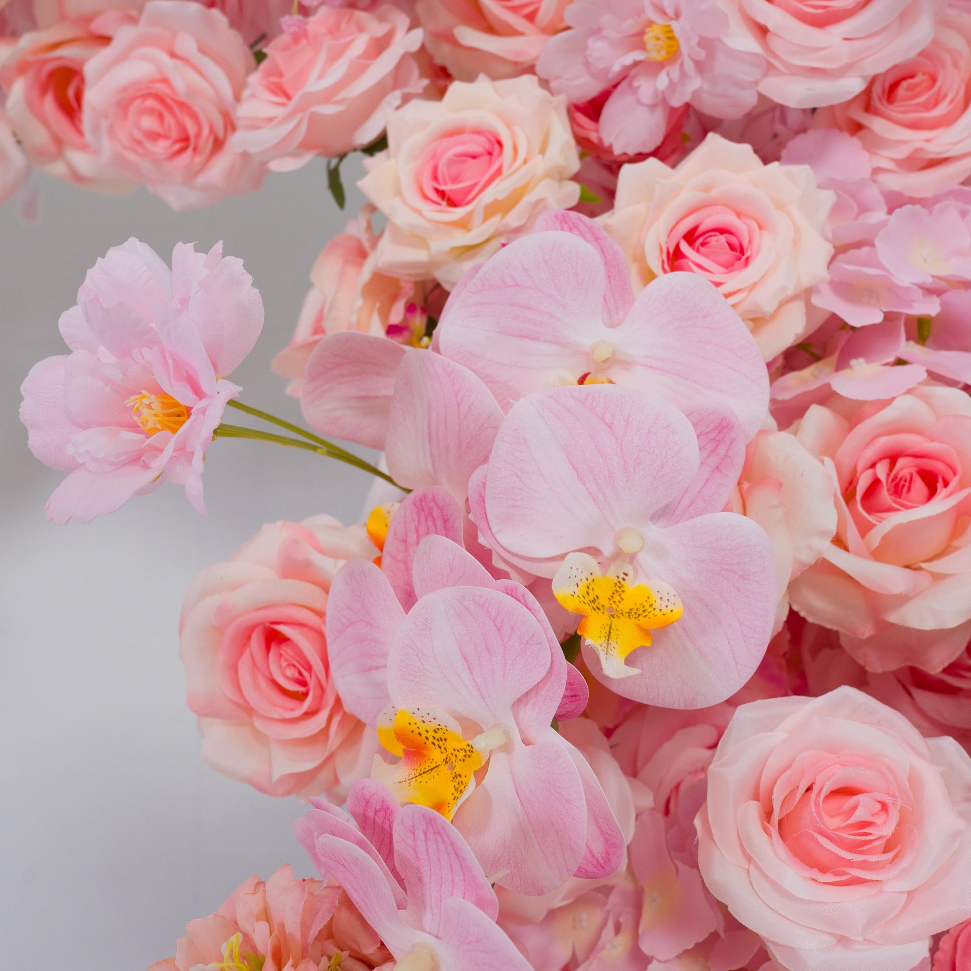 This backdrop features a lush assemblage of artificial flowers. Prominent are soft - pink roses with their classic, ruffled petals, and delicate pink orchids sporting vibrant yellow centers with black spots. There are also other pink blooms, adding to the variety.

The color scheme ranges from pale to deeper pinks, creating a harmonious and romantic palette. The overall style is elegant and dreamy, making it an ideal choice for romantic events like weddings or bridal showers. 