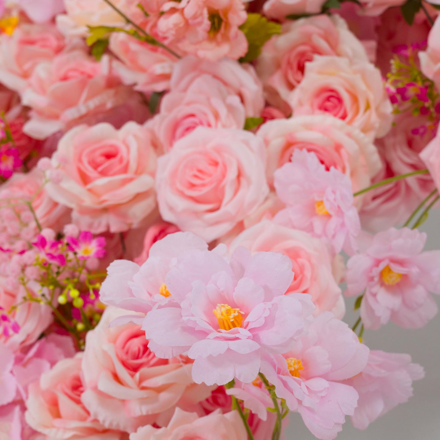 This backdrop features a charming arrangement of artificial flowers. It prominently includes various shades of pink roses, with their classic layered petals. There are also delicate pink blossoms with yellow centers,  and small clusters of vibrant pink filler flowers.

The color palette ranges from soft pastels to more vivid pinks, creating a warm and inviting look. The overall style is romantic and elegant, perfect for events like weddings, bridal showers, or other celebratory occasions. 