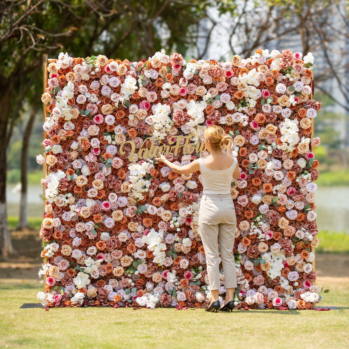 A woman with blonde hair, dressed in a sleeveless top and beige pants, stands in front of a lush floral backdrop. The backdrop is densely packed with roses and orchids in earthy tones, featuring the text "Valar Flowers", set in an outdoor park - like environment. 