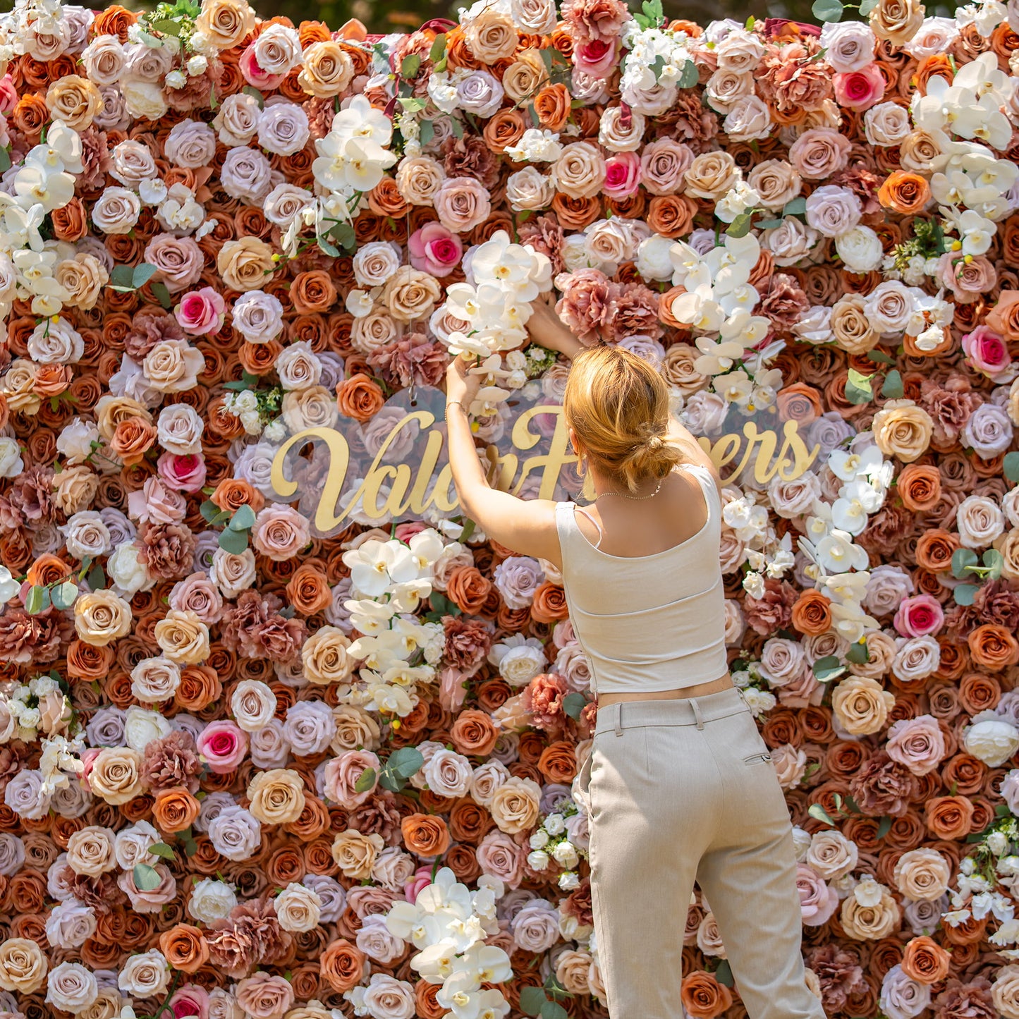 A woman with blonde hair, wearing a sleeveless top and beige pants, is adjusting flowers on a lush floral backdrop. The backdrop is densely packed with roses and orchids in earthy tones, and has the words "Valar Flowers" in gold. 