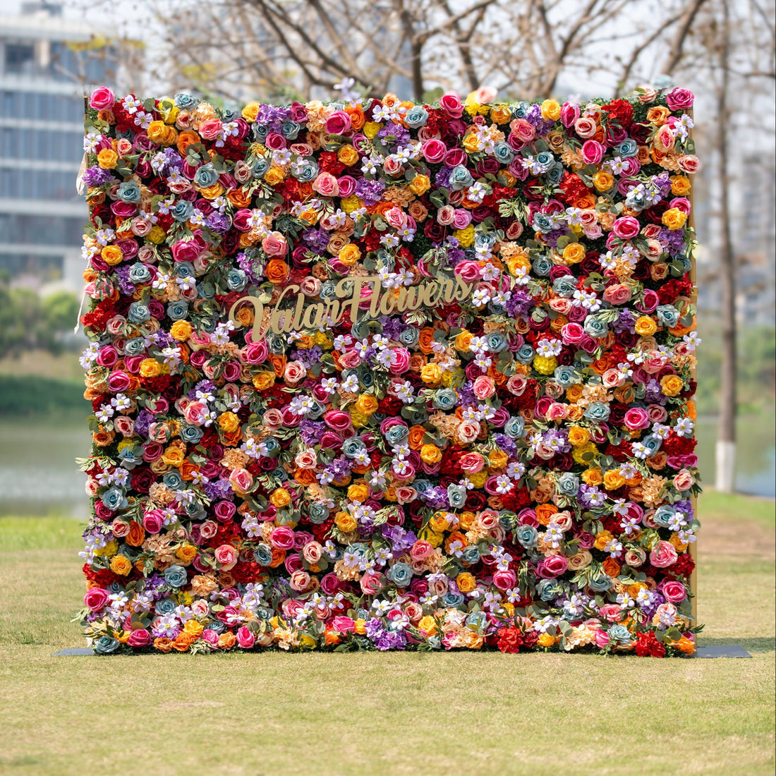 This image displays a vibrant floral backdrop brimming with multicolored roses in hues of red, orange, yellow, purple, and pink. Interspersed with smaller flowers and greenery, it features the text "Valar Flowers" in gold, ideal for festive outdoor events.