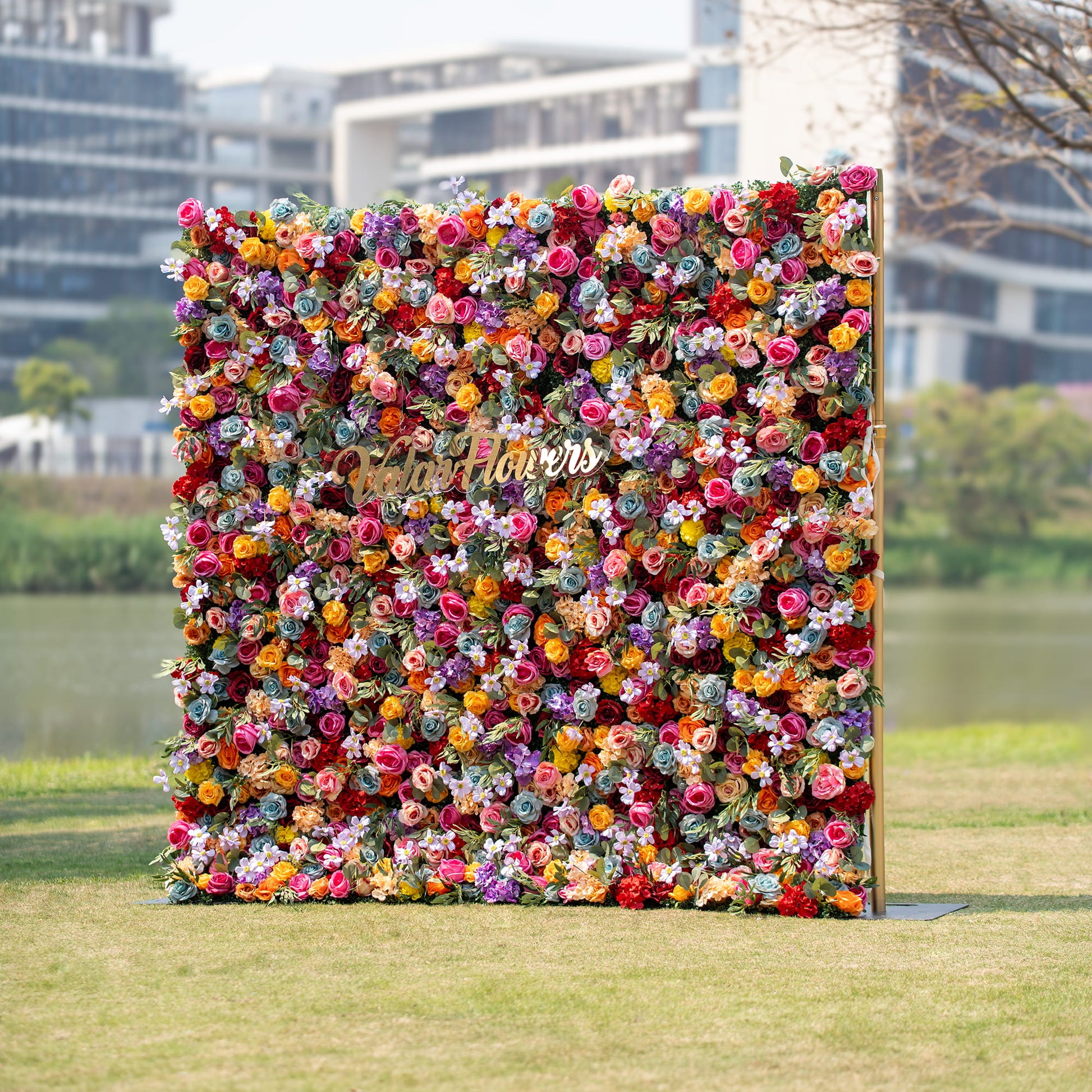 This image showcases a vibrant floral backdrop brimming with multicolored roses in hues of red, orange, yellow, purple, and pink. The lush arrangement features the text "Valar Flowers" in gold, set against an outdoor backdrop with buildings and a body of water. 