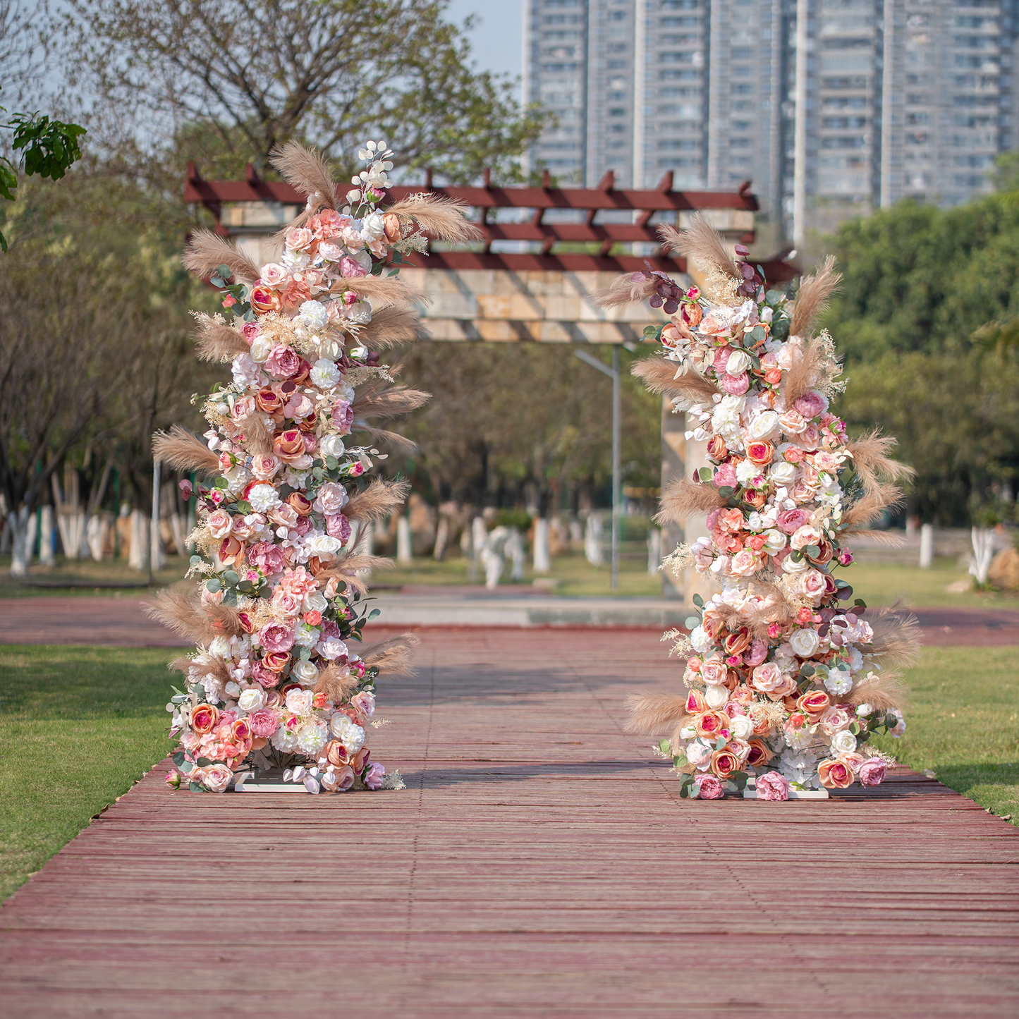 The photo showcases a beautifully adorned floral archway set on a wooden pathway in an outdoor park - like area. It's decorated with a mix of roses in soft pink, white, and peach hues, along with pampas grass, creating an elegant and inviting entrance for events like weddings. 