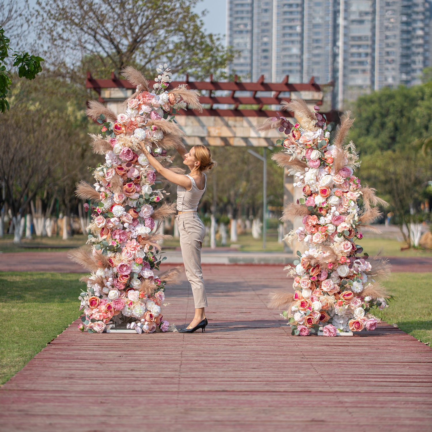 A woman with blonde hair, dressed in a sleeveless top and beige pants, is adjusting flowers on a floral archway. The arch is adorned with a mix of roses in pink, white, and peach hues, along with pampas grass, set on a wooden pathway in an outdoor park - like setting. 