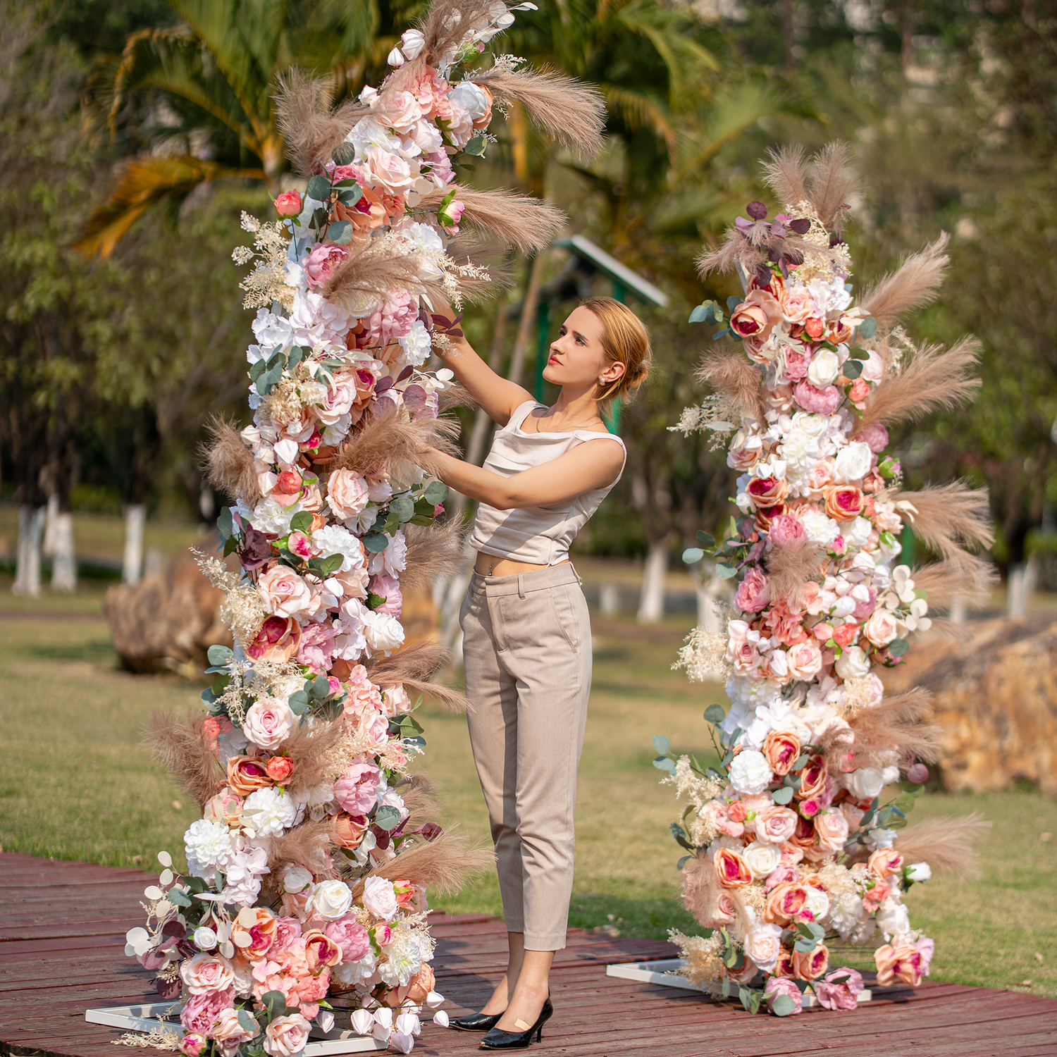 A woman in a sleeveless top and beige pants is arranging a floral arch. The arch is adorned with roses, peonies, and pampas grass in soft pink, white, and beige hues. Set outdoors with greenery in the background, it exudes a romantic, elegant style, perfect for weddings or garden parties. 