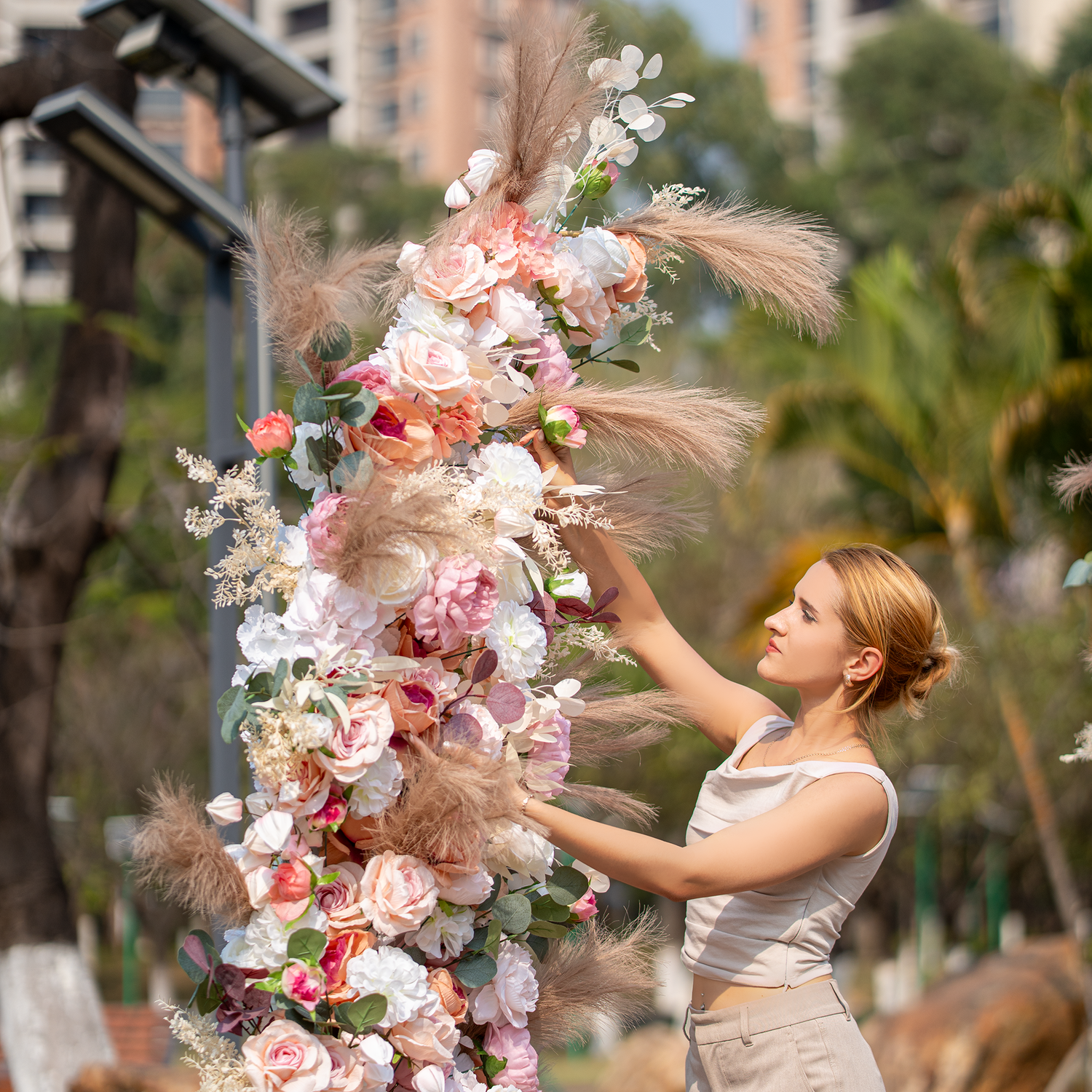 A woman in a sleeveless top and beige pants arranges a floral structure. It features roses, peonies, and pampas grass in soft pink, white, and beige tones. Set against an outdoor backdrop with greenery and buildings, it has a romantic, elegant style, ideal for weddings or garden parties. 