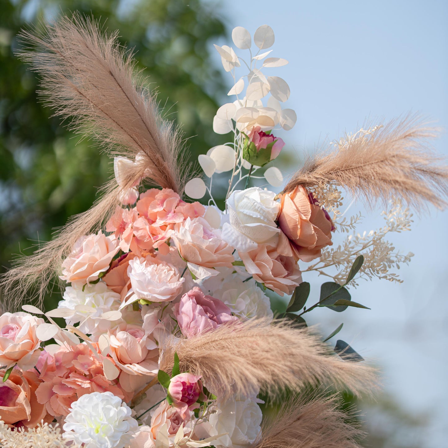 This photo showcases a close - up of a floral arrangement featuring roses and hydrangeas in soft pink, peach, and white hues, complemented by pampas grass and eucalyptus leaves. Set against a blurred outdoor green and blue sky backdrop, it has a romantic, elegant style, ideal for weddings or garden parties. 