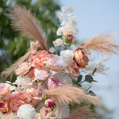 This photo showcases a close - up of a floral arrangement featuring roses and hydrangeas in soft pink, peach, and white hues, complemented by pampas grass and eucalyptus leaves. Set against a blurred outdoor green and blue sky backdrop, it has a romantic, elegant style, ideal for weddings or garden parties. 