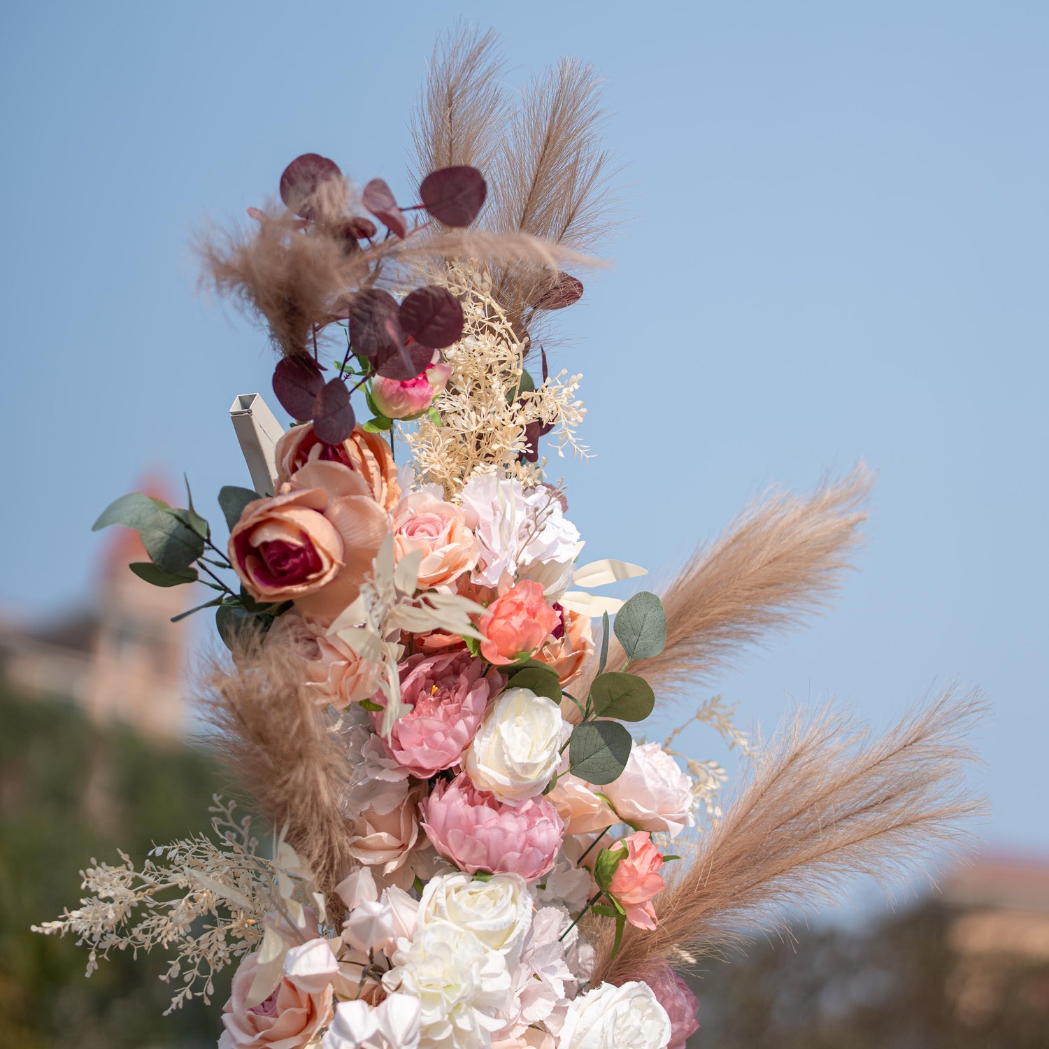 This photo shows a close - up of a floral arrangement with roses and peonies in soft pink, peach, and white tones. Pampas grass and eucalyptus leaves add texture. Set against a clear blue sky, it has a romantic, elegant style, perfect for weddings or upscale garden events. 