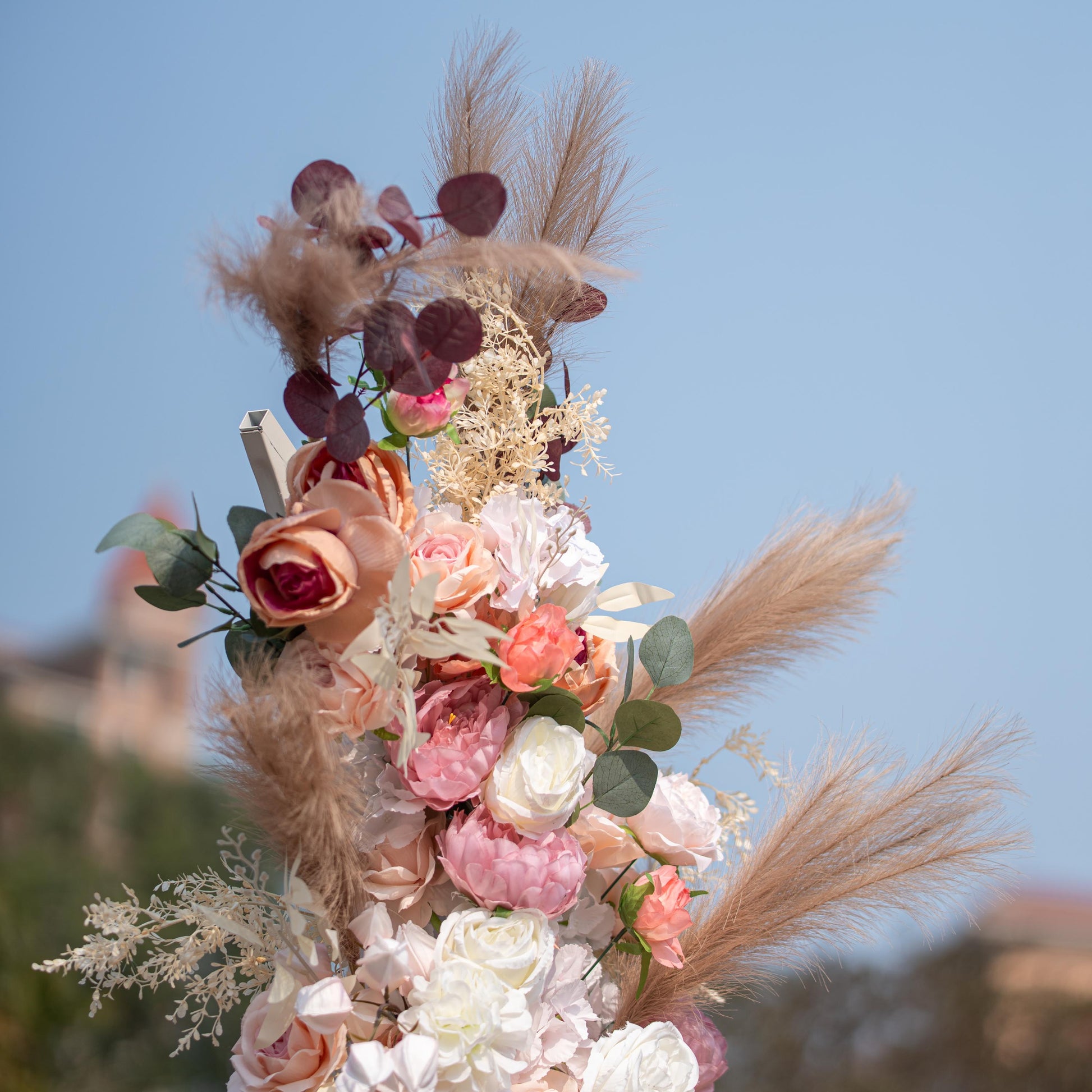 This photo shows a close - up of a floral arrangement with roses and peonies in soft pink, peach, and white tones. Pampas grass and eucalyptus leaves add texture. Set against a clear blue sky, it has a romantic, elegant style, perfect for weddings or upscale garden events. 