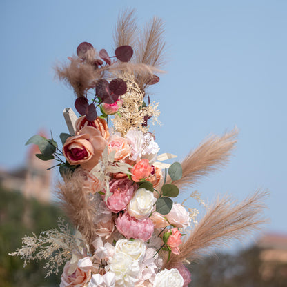 This photo shows a close - up of a floral arrangement with roses and peonies in soft pink, peach, and white tones. Pampas grass and eucalyptus leaves add texture. Set against a clear blue sky, it has a romantic, elegant style, perfect for weddings or upscale garden events. 