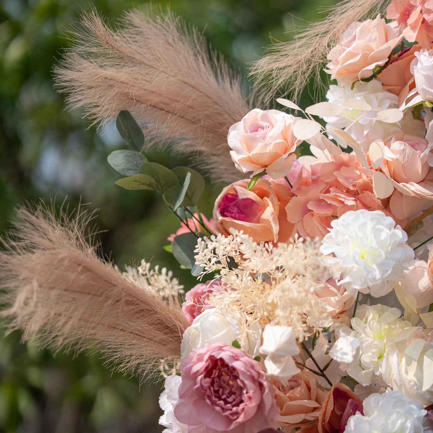 This close - up photo features a floral arrangement with roses in soft pink and peach tones, along with white blooms. Pampas grass and eucalyptus leaves add texture. Set against a blurred green outdoor backdrop, it has a romantic, elegant style, perfect for weddings or garden - themed events. 