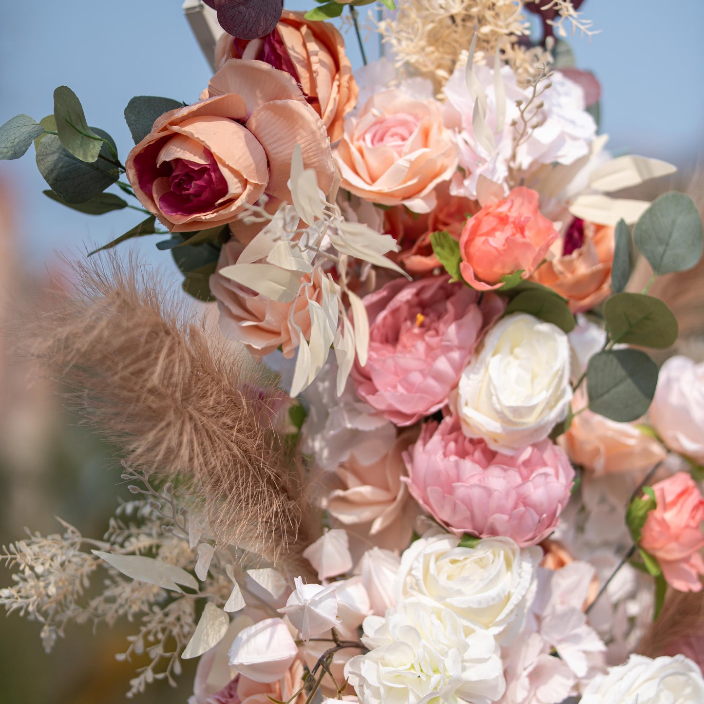 This photo captures a lush floral arrangement featuring roses and peonies in soft pink, peach, and white hues. Pampas grass and eucalyptus leaves add texture. Set against a clear blue sky, it exudes a romantic, elegant style, ideal for weddings or garden - themed events. 
