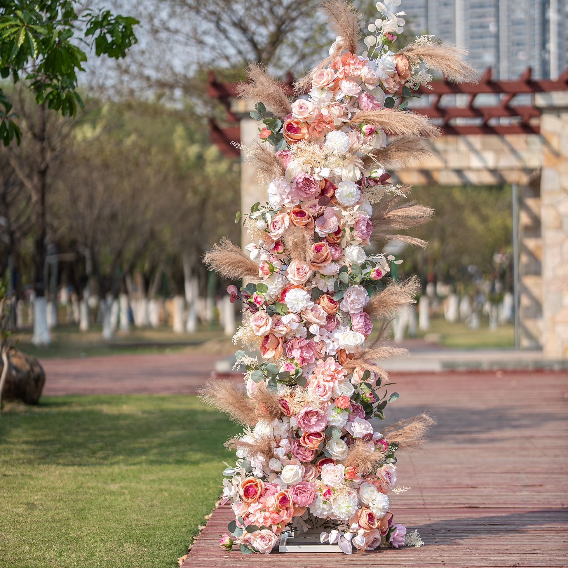 This photo features a tall floral arrangement, likely part of an arch, brimming with roses and peonies in soft pink, peach, and white shades. Pampas grass and eucalyptus leaves add texture. Set outdoors with greenery and a structure in the background, it has a romantic, elegant style, perfect for weddings. 