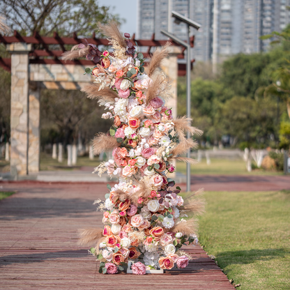 This photo showcases a tall, elaborate floral arrangement, likely part of an arch, bursting with roses and peonies in soft pink, peach, and white tones. Pampas grass and eucalyptus leaves add texture. Set outdoors with greenery and city buildings in the background, it has a romantic, elegant style, perfect for weddings or upscale garden events. 