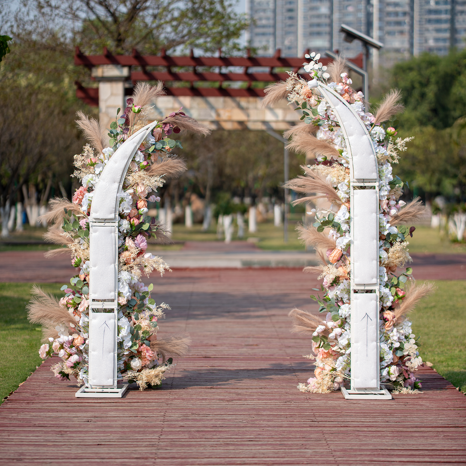 This photo features a floral - adorned archway with roses and peonies in soft pink, peach, and white hues. Pampas grass and eucalyptus leaves add texture. Set outdoors on a wooden path with greenery and a pergola in the background, it has a romantic, elegant style, ideal for weddings or garden parties. 