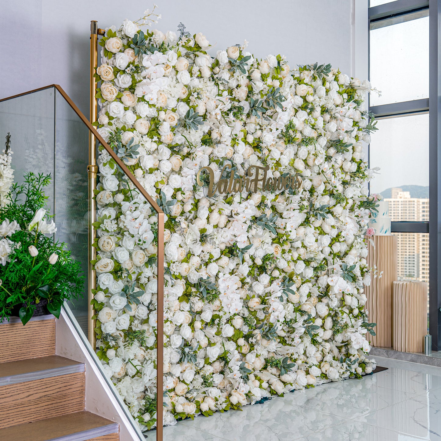 This backdrop is a lush, elegant display of white artificial flowers mixed with green foliage, creating a soft, romantic look. The golden "Valar Flowers" sign adds a touch of luxury, making it perfect for weddings or upscale events. 