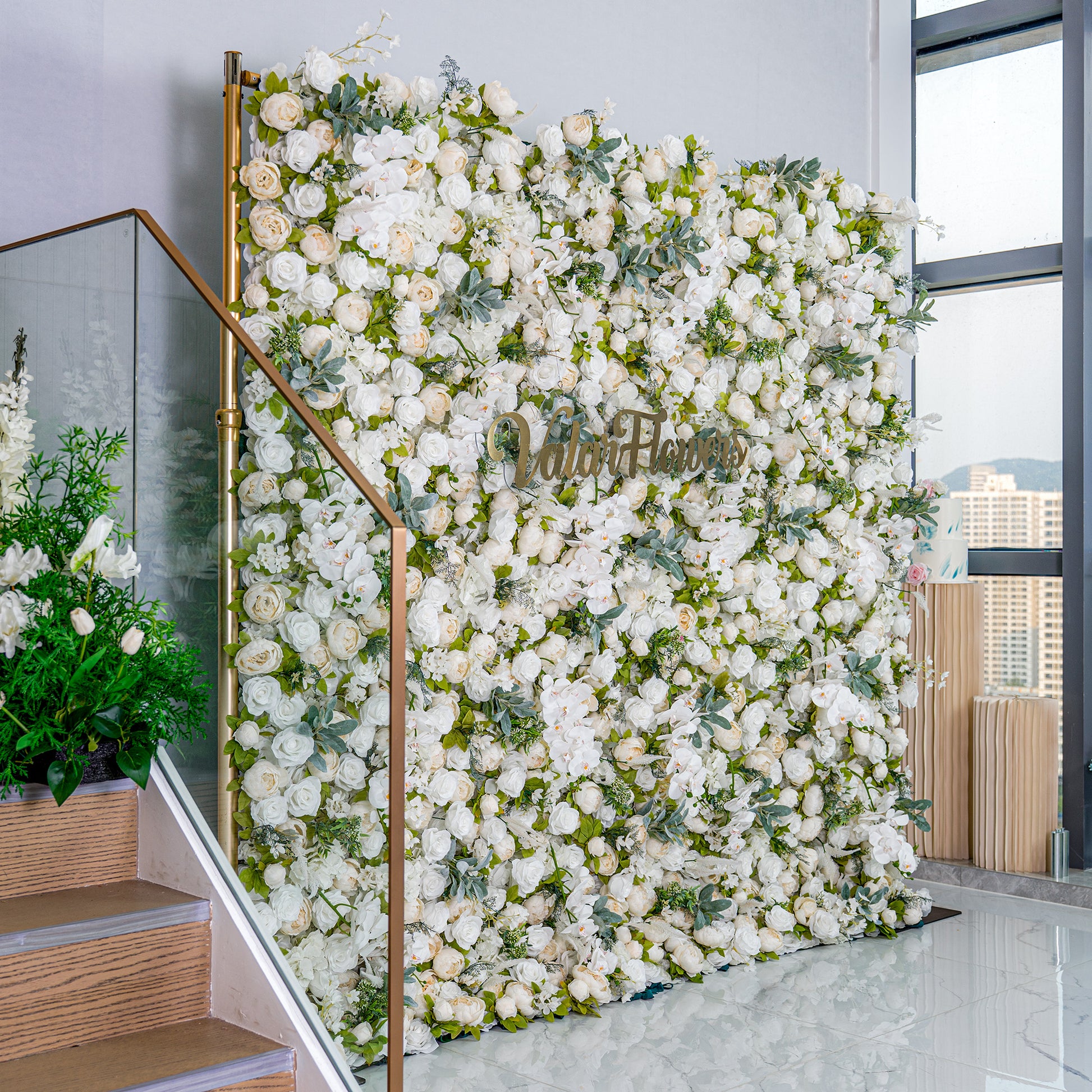 This backdrop is a lush, elegant display of white artificial flowers mixed with green foliage, creating a soft, romantic look. The golden "Valar Flowers" sign adds a touch of luxury, making it perfect for weddings or upscale events. 