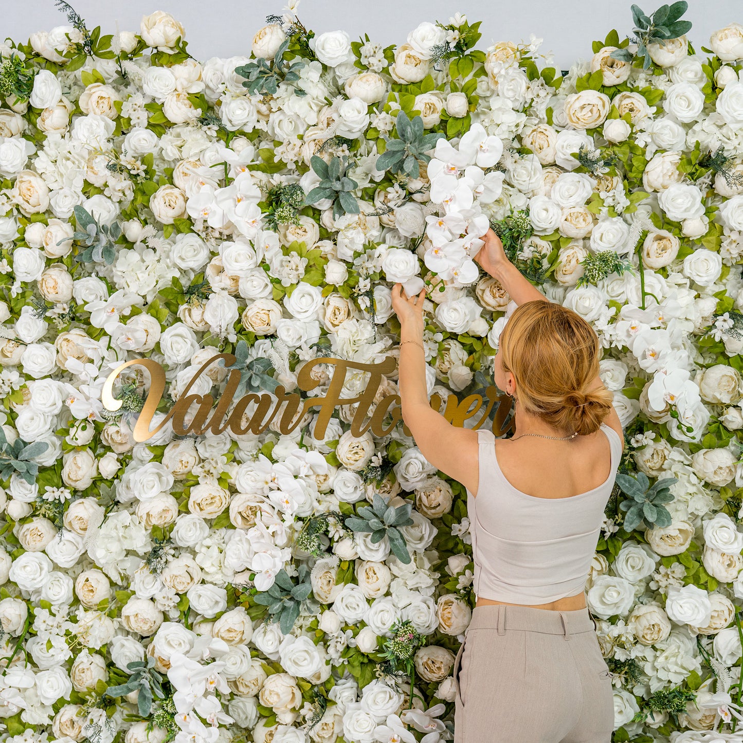 A woman with blonde hair, dressed in a light - colored top and pants, is arranging white flowers on a lush floral backdrop. The backdrop features the text "Valar Flowers" in gold, with numerous white blooms and green foliage. 