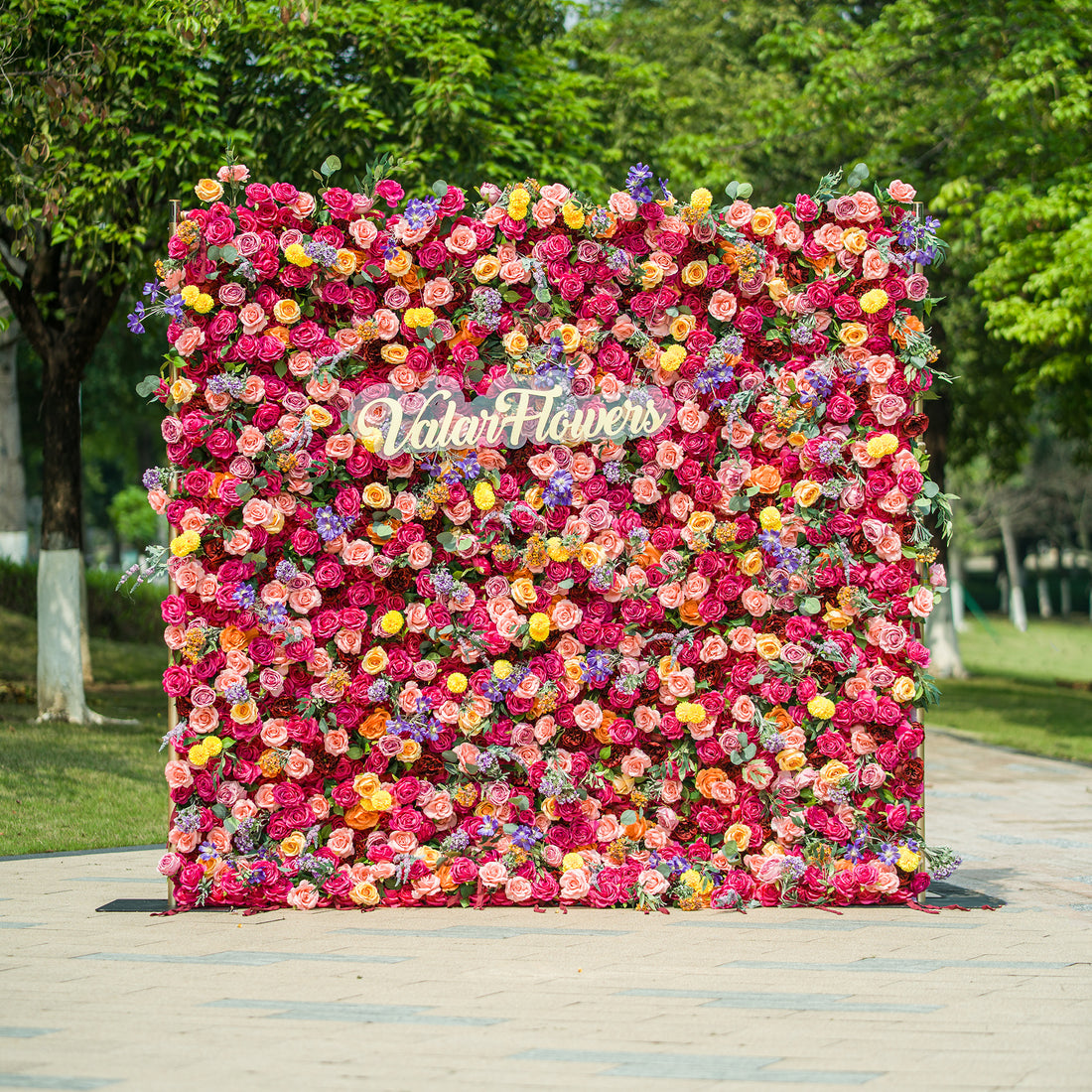 This vibrant floral wall by ValarFlowers is a stunning display of color and craftsmanship. It features a dense arrangement of artificial flowers in a wide range of hues, including pink, red, yellow, purple, and orange. The variety of blooms, such as roses, daisies, and other floral shapes, creates a lush and textured appearance. The “ValarFlowers” logo is prominently displayed in the center, adding a branded touch. Set against an outdoor backdrop with greenery, this floral wall is perfect for events, photo 