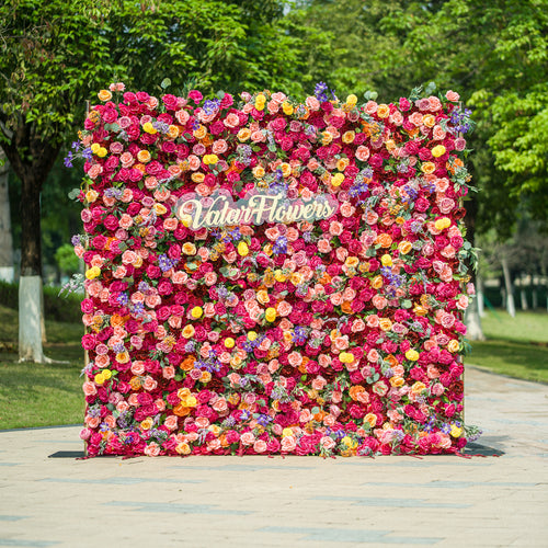 This vibrant floral wall by ValarFlowers is a stunning display of color and craftsmanship. It features a dense arrangement of artificial flowers in a wide range of hues, including pink, red, yellow, purple, and orange. The variety of blooms, such as roses, daisies, and other floral shapes, creates a lush and textured appearance. The “ValarFlowers” logo is prominently displayed in the center, adding a branded touch. Set against an outdoor backdrop with greenery, this floral wall is perfect for events, photo 