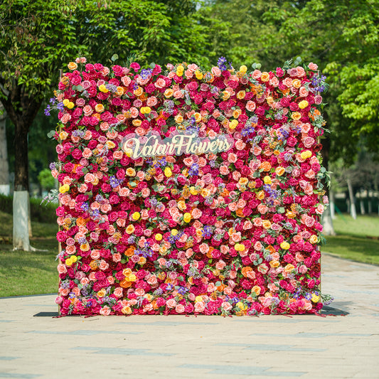 This vibrant floral wall by ValarFlowers is a stunning display of color and craftsmanship. It features a dense arrangement of artificial flowers in a wide range of hues, including pink, red, yellow, purple, and orange. The variety of blooms, such as roses, daisies, and other floral shapes, creates a lush and textured appearance. The “ValarFlowers” logo is prominently displayed in the center, adding a branded touch. Set against an outdoor backdrop with greenery, this floral wall is perfect for events, photo 