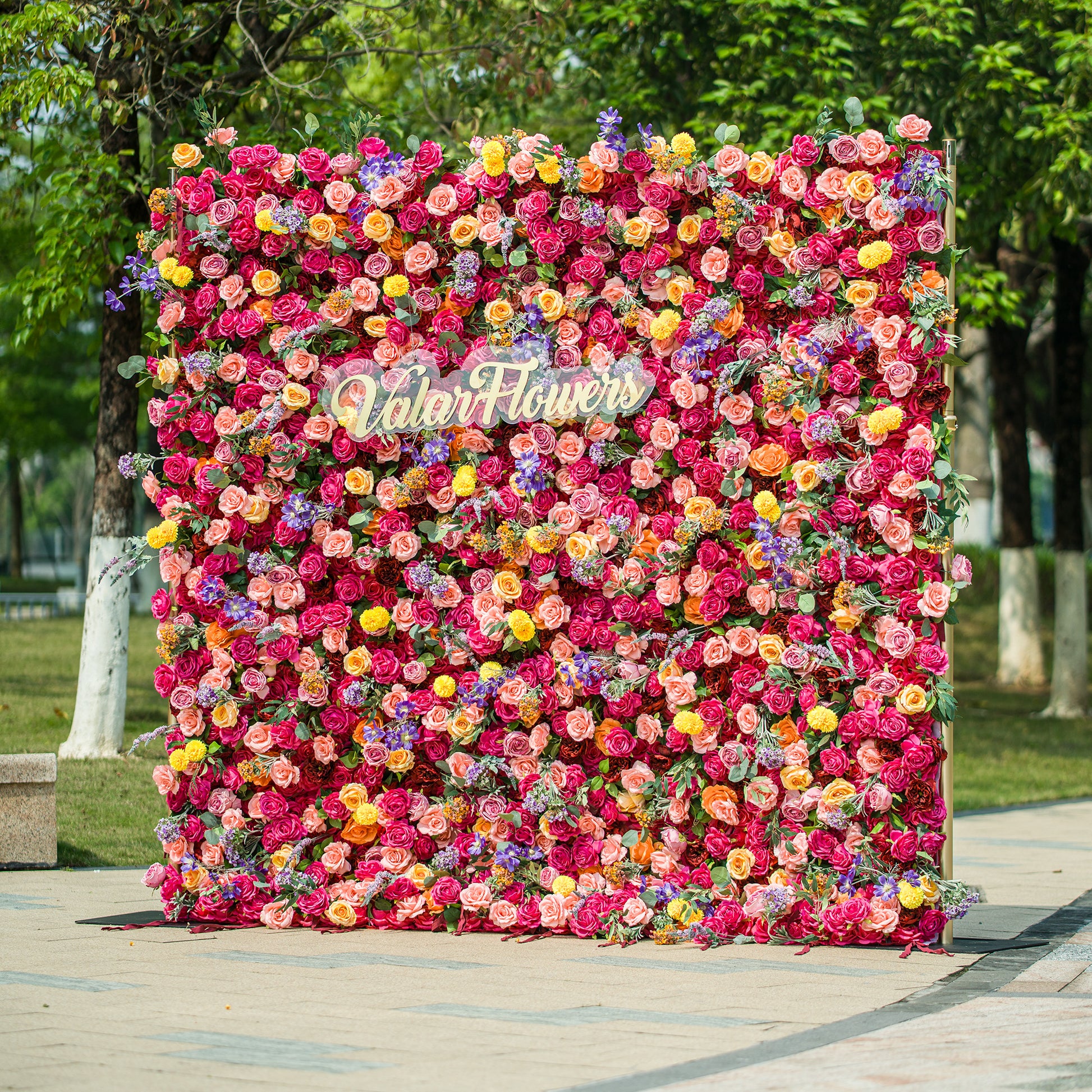 A vibrant floral wall by ValarFlowers bursts with color! Dense with roses, daisies, lavender, and more in shades of pink, red, yellow, purple—each bloom meticulously arranged. The gold “ValarFlowers” logo stands out at the center, set against lush green foliage in a park - like outdoor setting. Perfect for events, it creates a lively, Instagram - worthy backdrop full of texture and charm. 