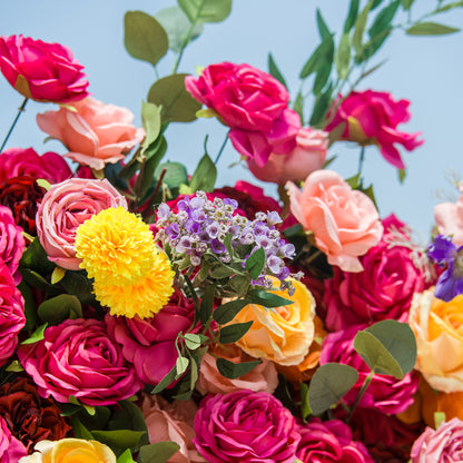 A vibrant floral arrangement bursts with color against a bright sky. Deep pink roses dominate, interspersed with soft peach and yellow blooms. A cluster of purple flowers and fluffy yellow blossoms add contrast, while eucalyptus leaves provide fresh greenery. The dense, varied blooms create a lively, eye-catching display, perfect for celebrations or decorative focal points. 