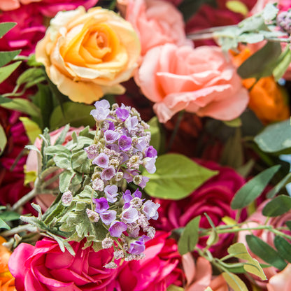 A close - up of a vibrant floral arrangement showcases a mix of blooms. At the center, delicate purple flowers with tiny, textured petals and soft green leaves stand out. Surrounding them are large, lush roses in shades of pink, peach, and yellow. The rich colors and varied textures create a visually appealing and lively composition, perfect for adding a touch of natural beauty to any setting. 