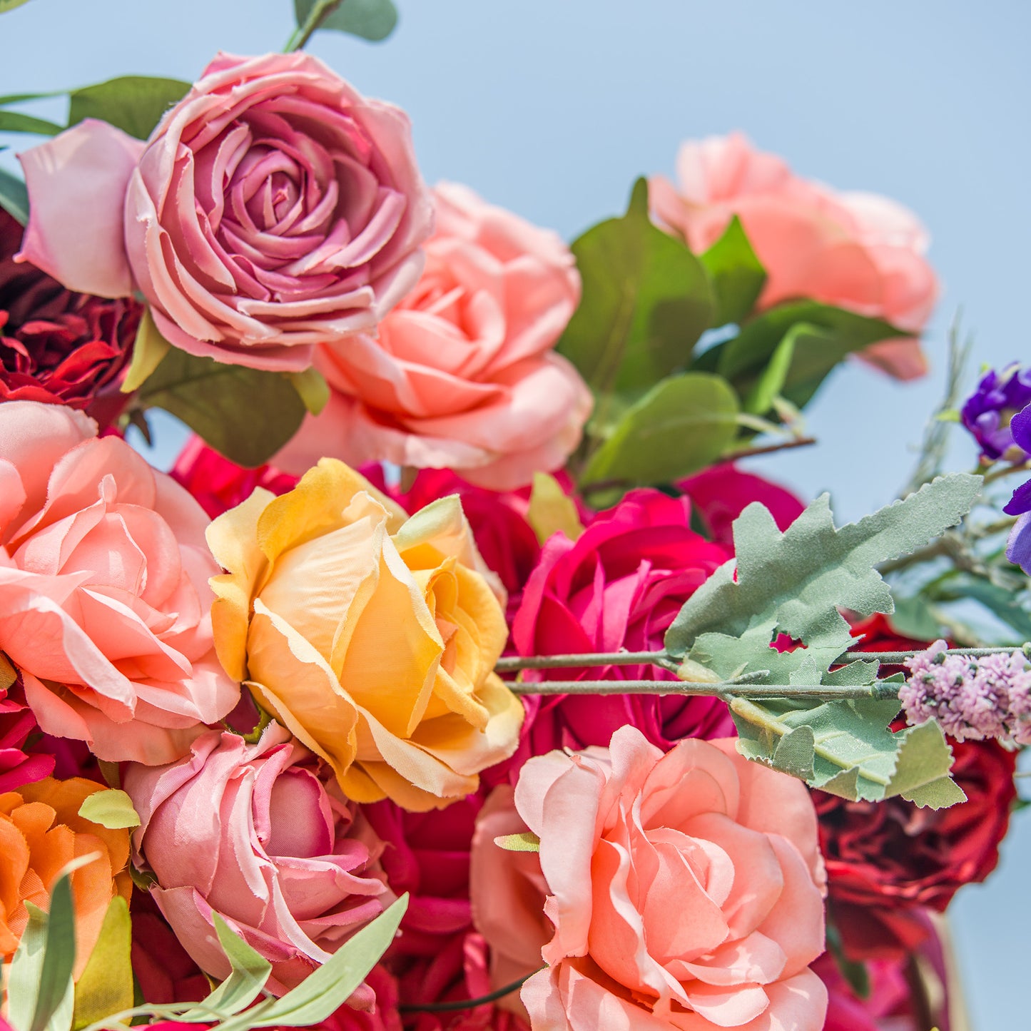 A vibrant floral arrangement bursts with color! Soft pink, peach, and magenta roses mix with a bright yellow rose. Green foliage and a hint of purple blossoms add texture. The clear sky backdrop enhances the vivid hues, creating a lively, eye - catching display perfect for celebrations or decorative focal points. 