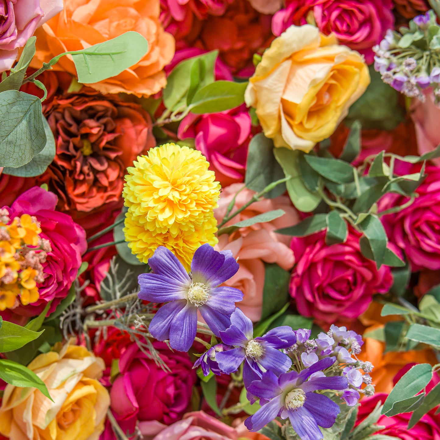 A vibrant floral arrangement showcases a rich tapestry of colors and textures. Bold pink and orange roses form a lush backdrop, interspersed with bright yellow pom - pom flowers and delicate purple blossoms. Green foliage weaves through, adding freshness. The dense, varied blooms create a lively, eye - catching display, perfect for adding a burst of natural beauty to any setting. 