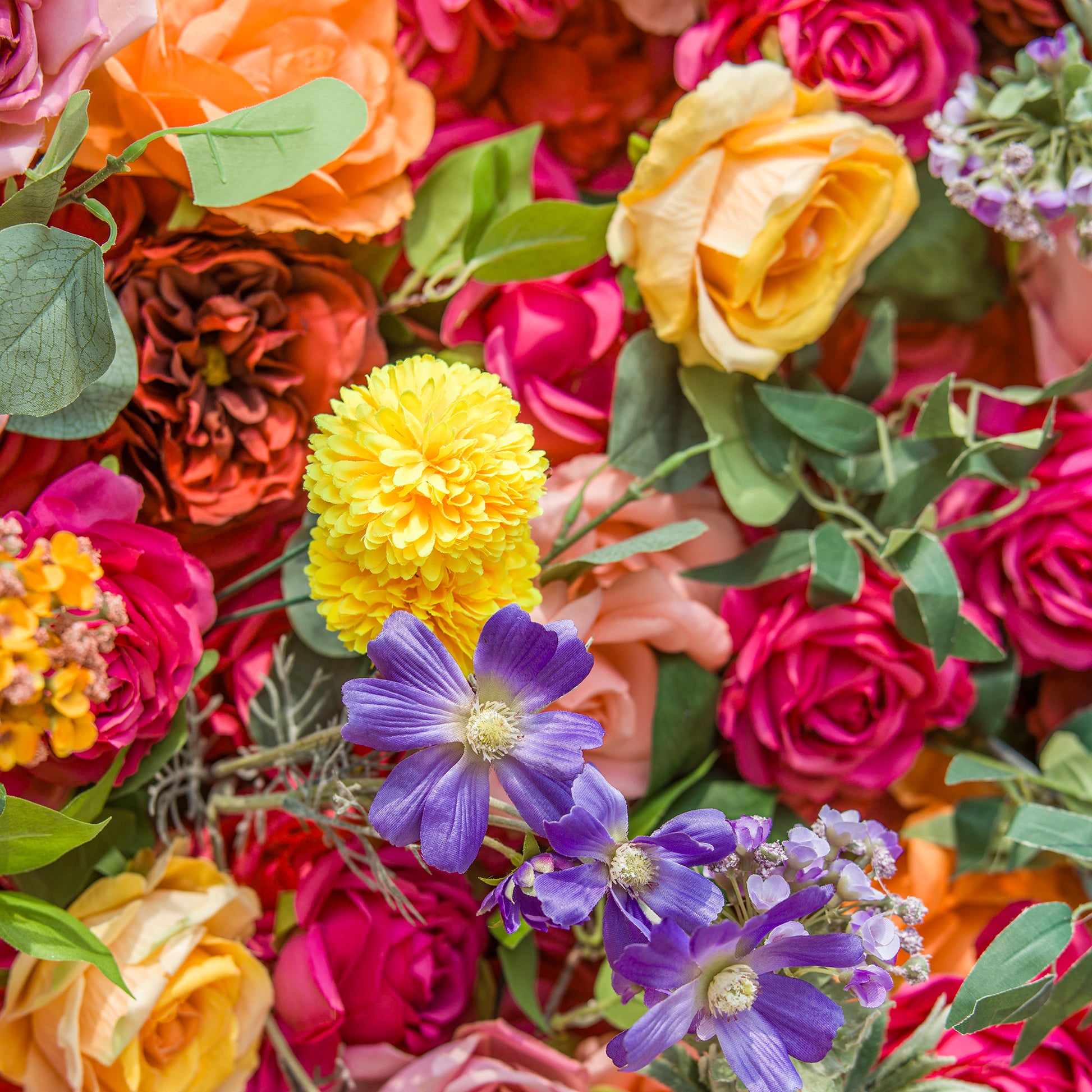A vibrant floral arrangement showcases a rich tapestry of colors and textures. Bold pink and orange roses form a lush backdrop, interspersed with bright yellow pom - pom flowers and delicate purple blossoms. Green foliage weaves through, adding freshness. The dense, varied blooms create a lively, eye - catching display, perfect for adding a burst of natural beauty to any setting. 