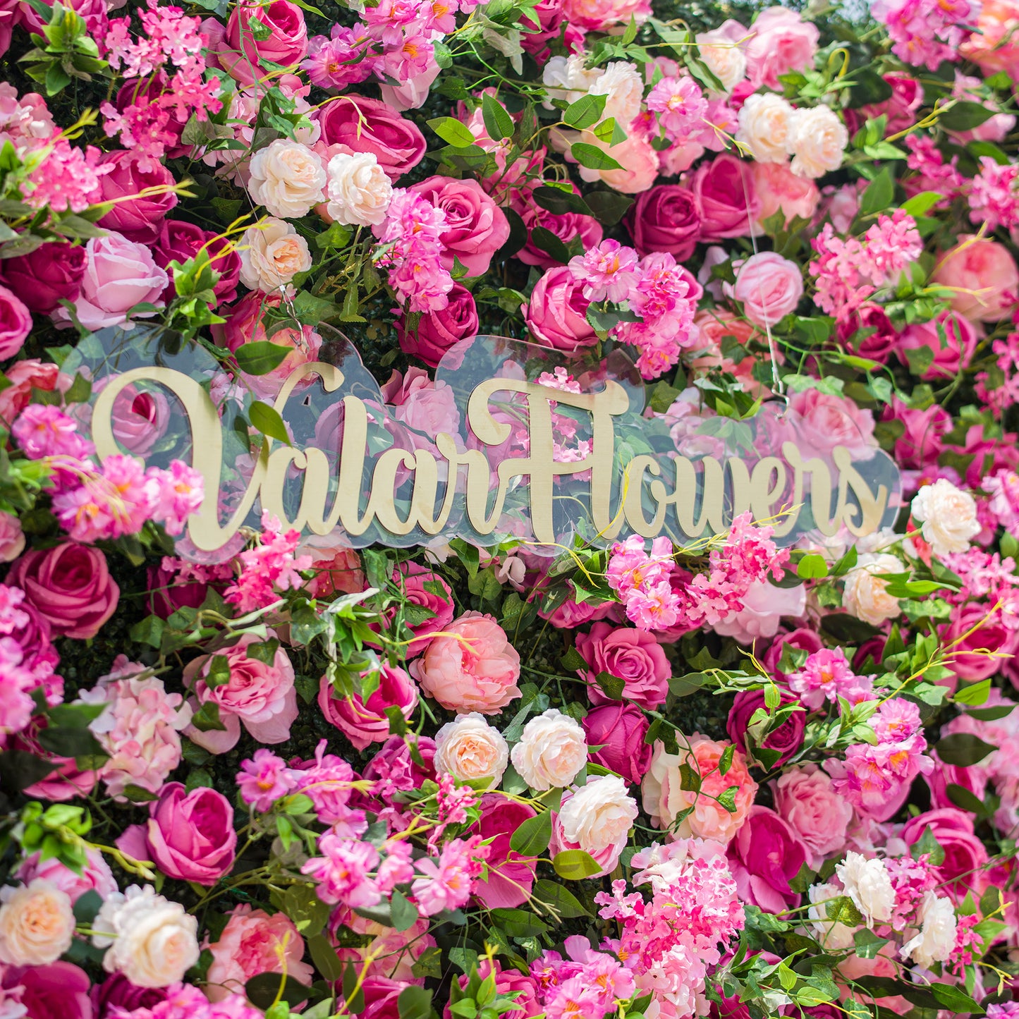 This photo showcases a lush and vibrant floral backdrop densely packed with an array of pink and white roses, along with other assorted pink flowers, all interspersed with rich green foliage. The text "ValarFlowers" in elegant golden lettering stands out prominently. The overall scene exudes a lively, romantic charm, making it ideal for events like garden weddings or spring festivals. 